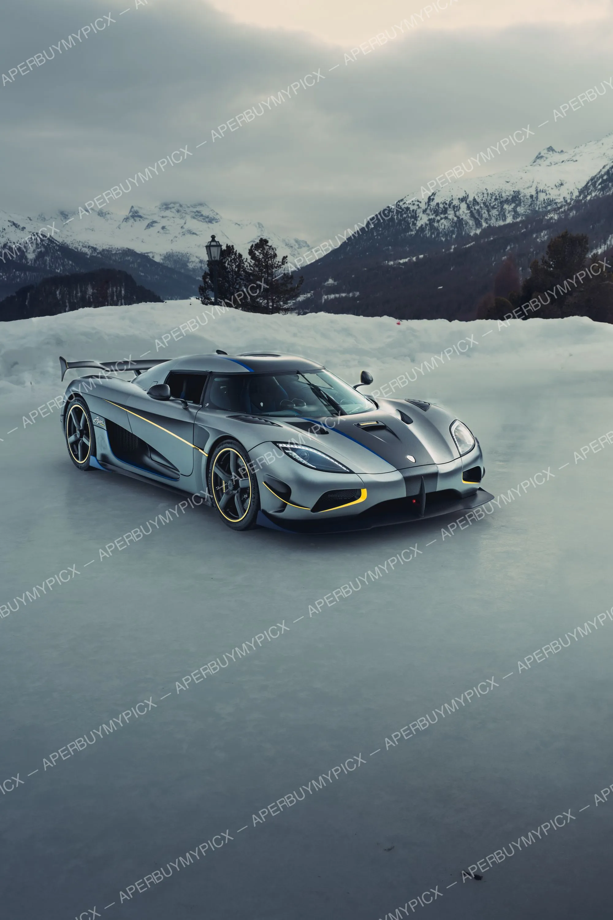 Koenigsegg Agera grise à liserés jaunes et bleus stationnée sur un lac gelé de St. Moritz, montagnes enneigées et ciel nuageux en arrière-plan.