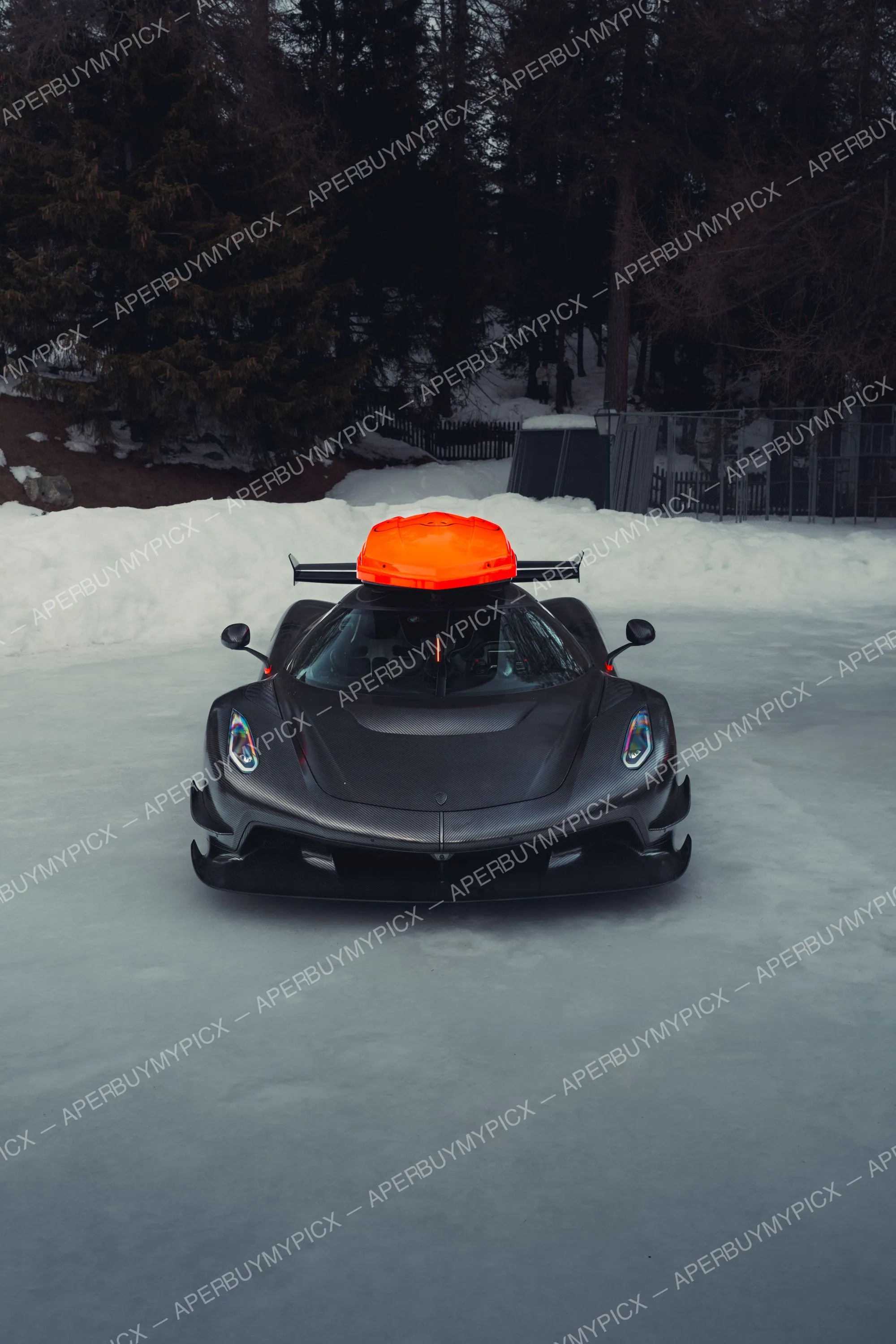 Hypercar Koenigsegg en fibre de carbone avec coffre de toit orange, garée sur un lac gelé enneigé bordé de sapins à St. Moritz.