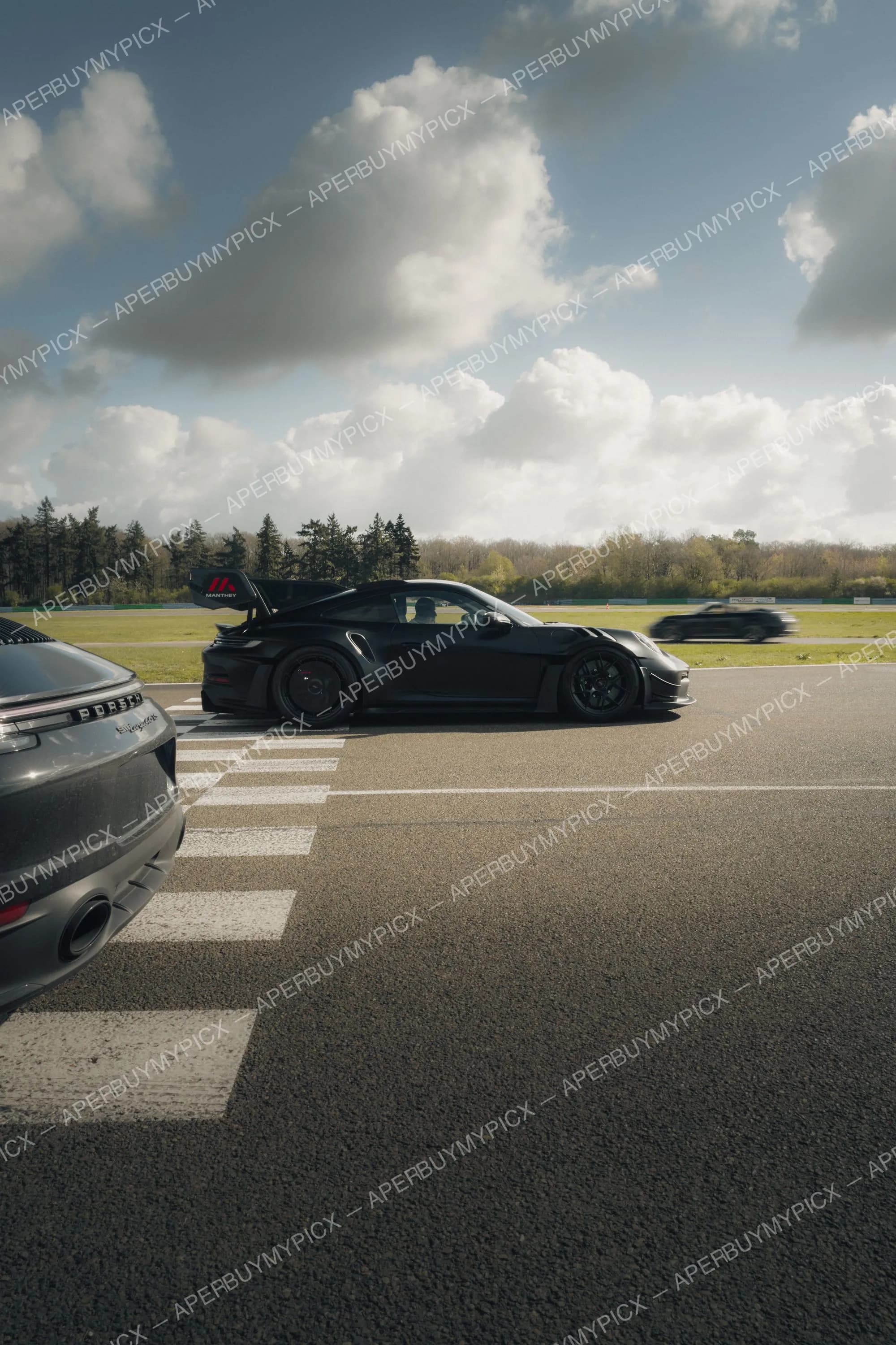 Porsche 911 GT3 RS noire avec aileron Manthey sur une piste de circuit, une Porsche grise au premier plan et une voiture floue en arrière-plan sous un ciel nuageux.