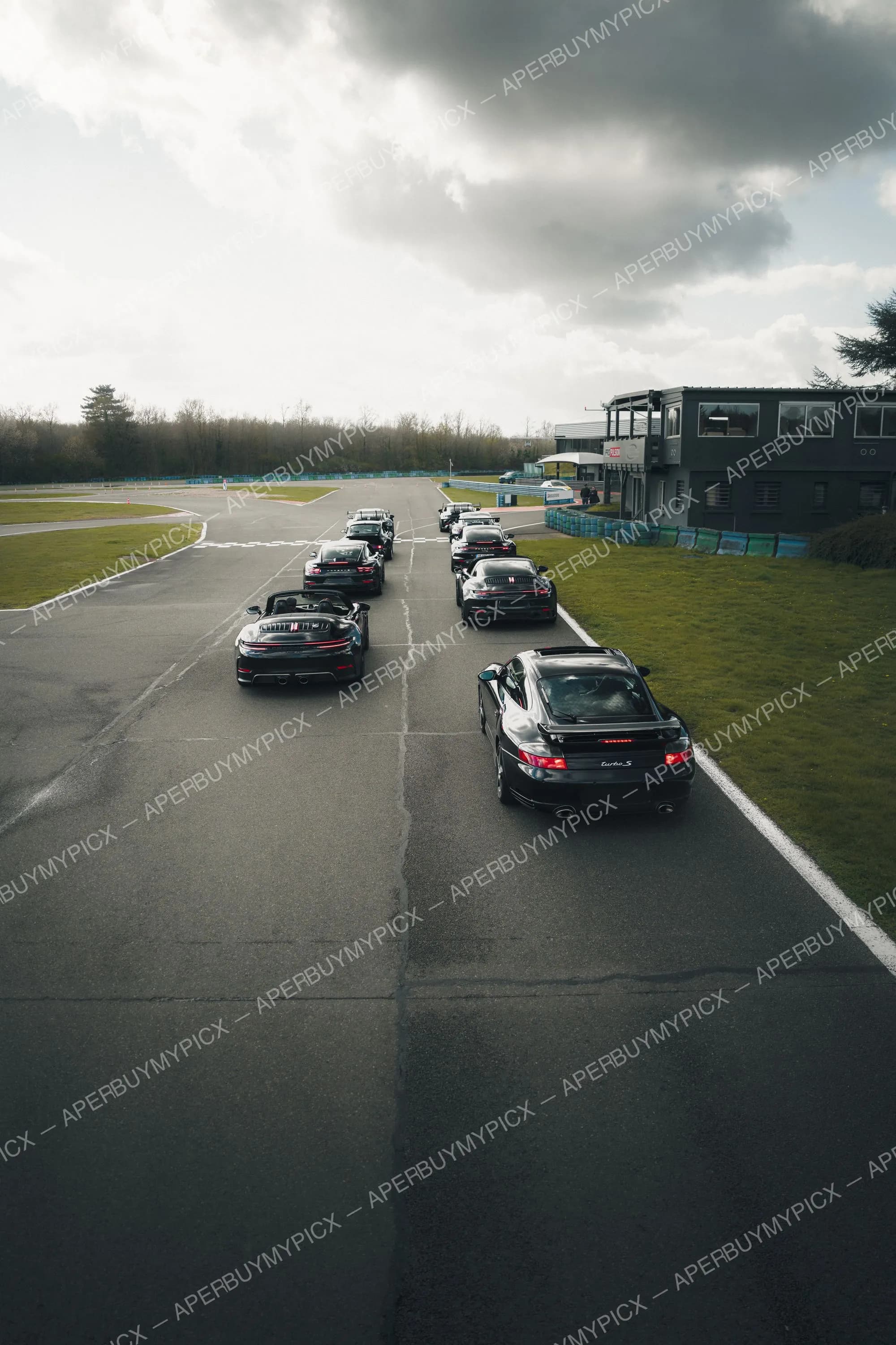 Groupe de Porsche 911 noires, dont une 996 Turbo S et un cabriolet, alignées sur la voie des stands d'un circuit sous un ciel gris nuageux, avec bâtiment de direction de course à droite.