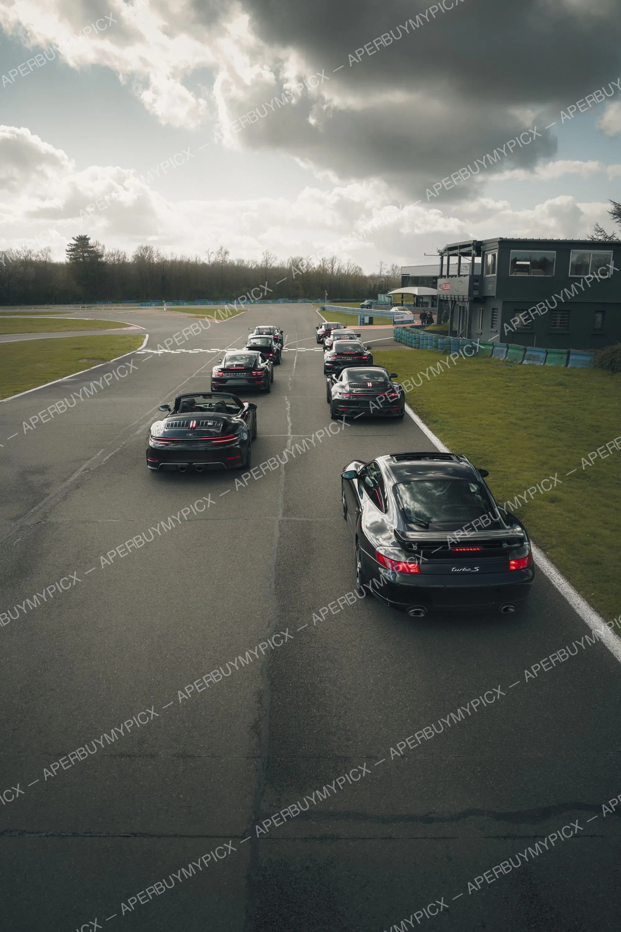 Rassemblement de Porsche 911 noires, dont un cabriolet et une Turbo S, alignées sur une piste de circuit sous un ciel nuageux, avec bâtiment de contrôle à droite.