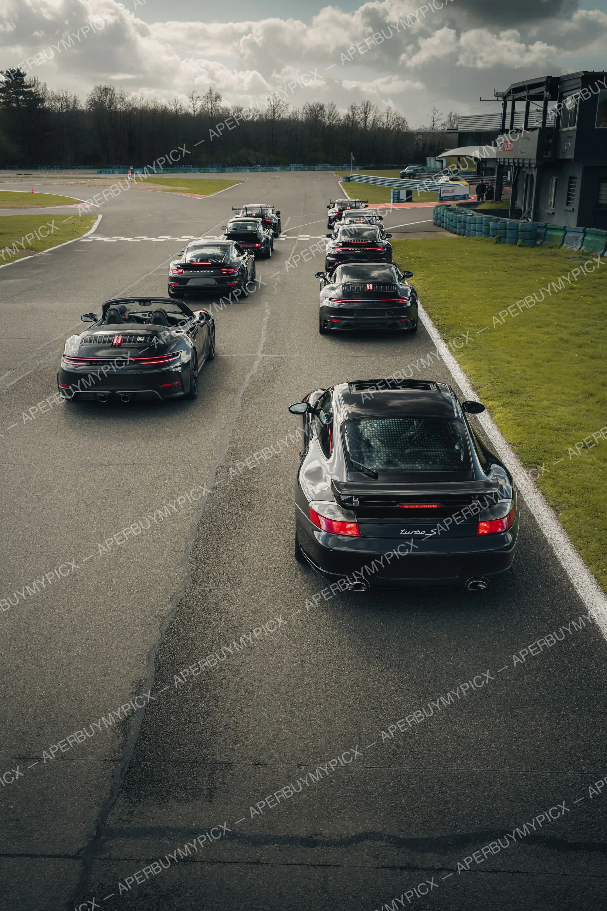 Rassemblement de plusieurs Porsche 911 noires alignées sur deux files sur une piste de circuit, avec au premier plan une 911 Turbo S, un cabriolet et des berlines sportives sous un ciel nuageux, bâtiment de stand à droite.