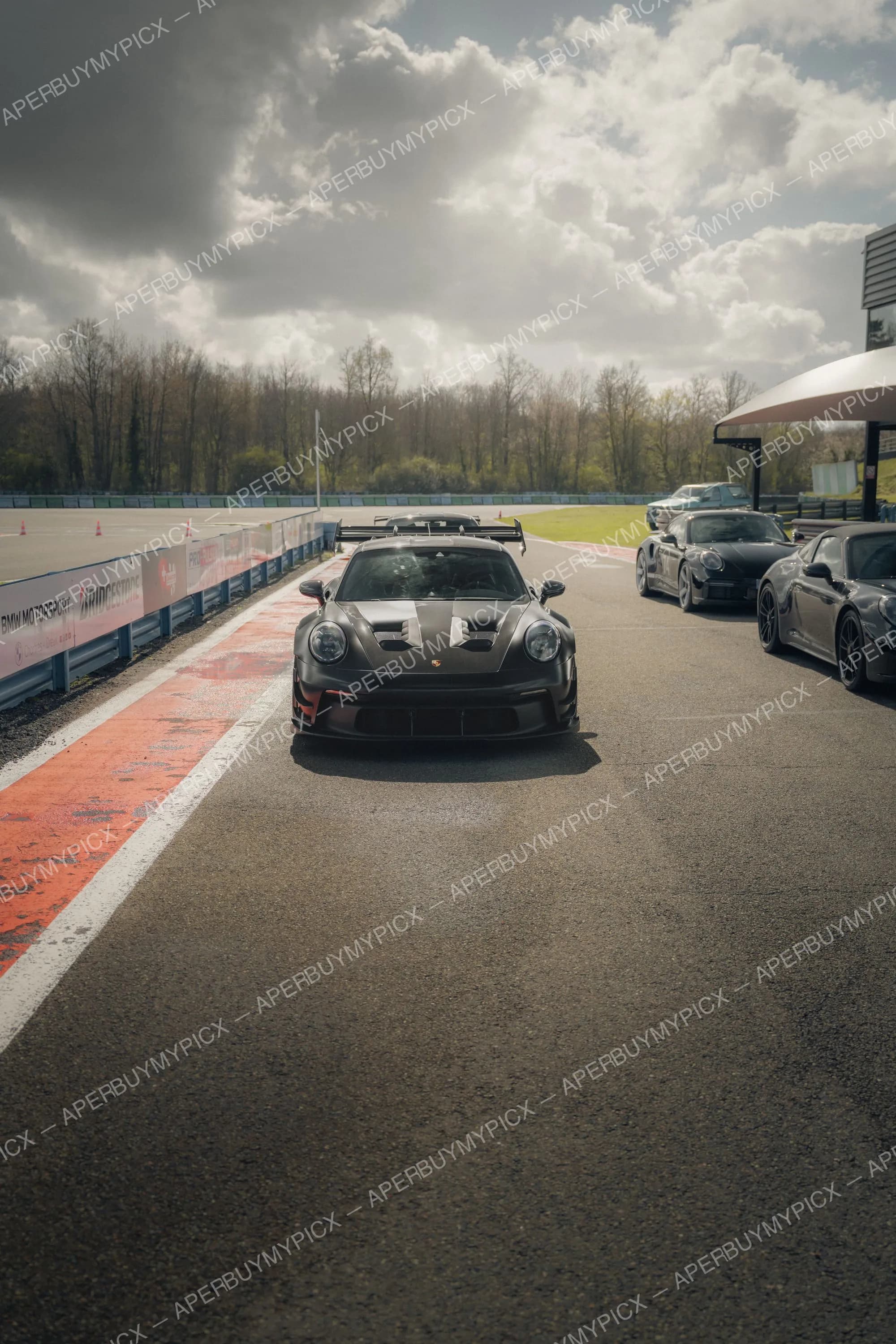 Porsche 911 GT3 RS noire vue de face dans la voie des stands d'un circuit, avec aileron arrière et capot à prises d'air, suivie de plusieurs Porsche 911 sombres sous un ciel nuageux.