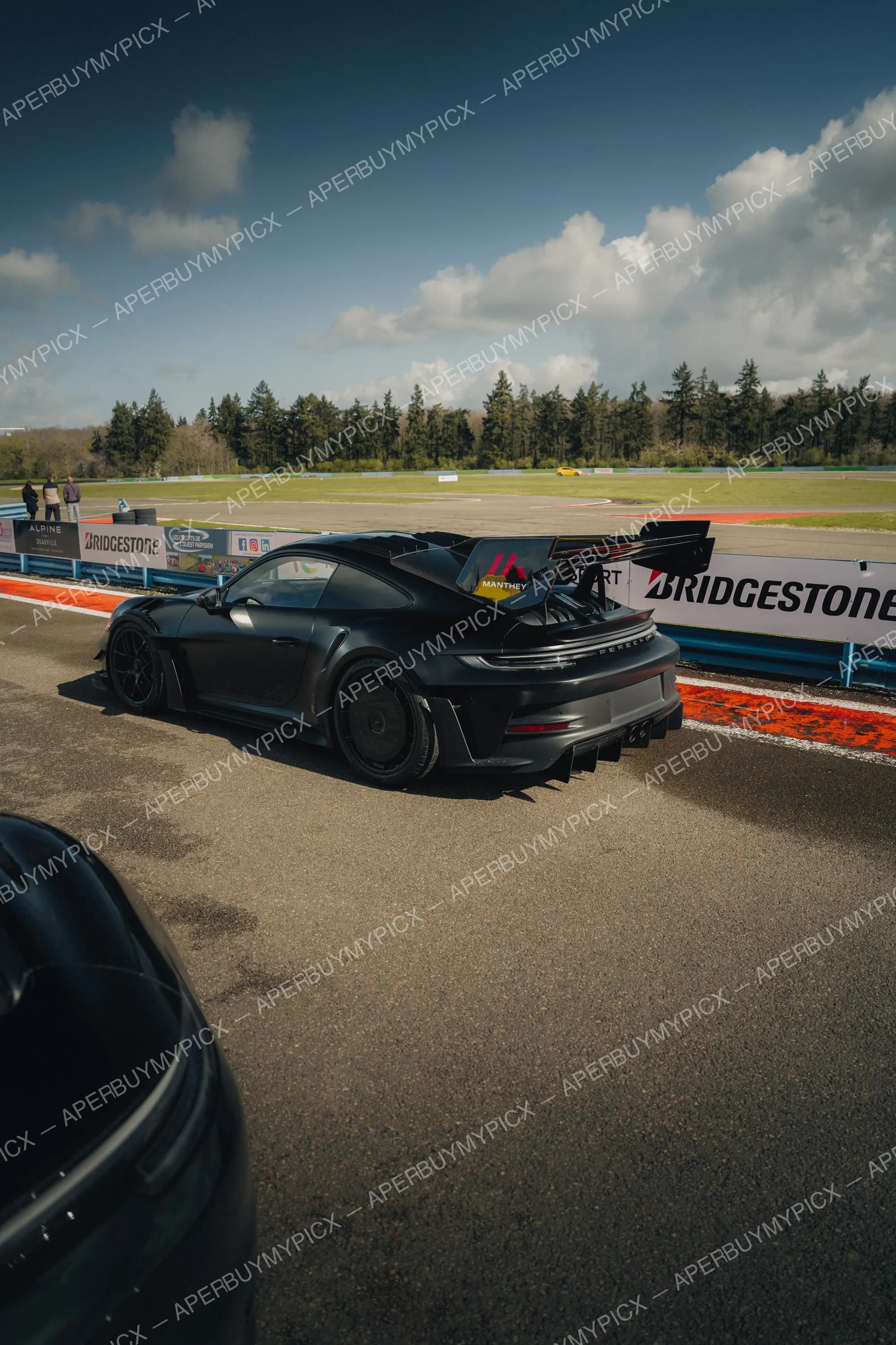 Porsche 911 GT3 noire mate avec kit Manthey et aileron arrière imposant, garée sur la piste devant des panneaux Bridgestone sous un ciel nuageux