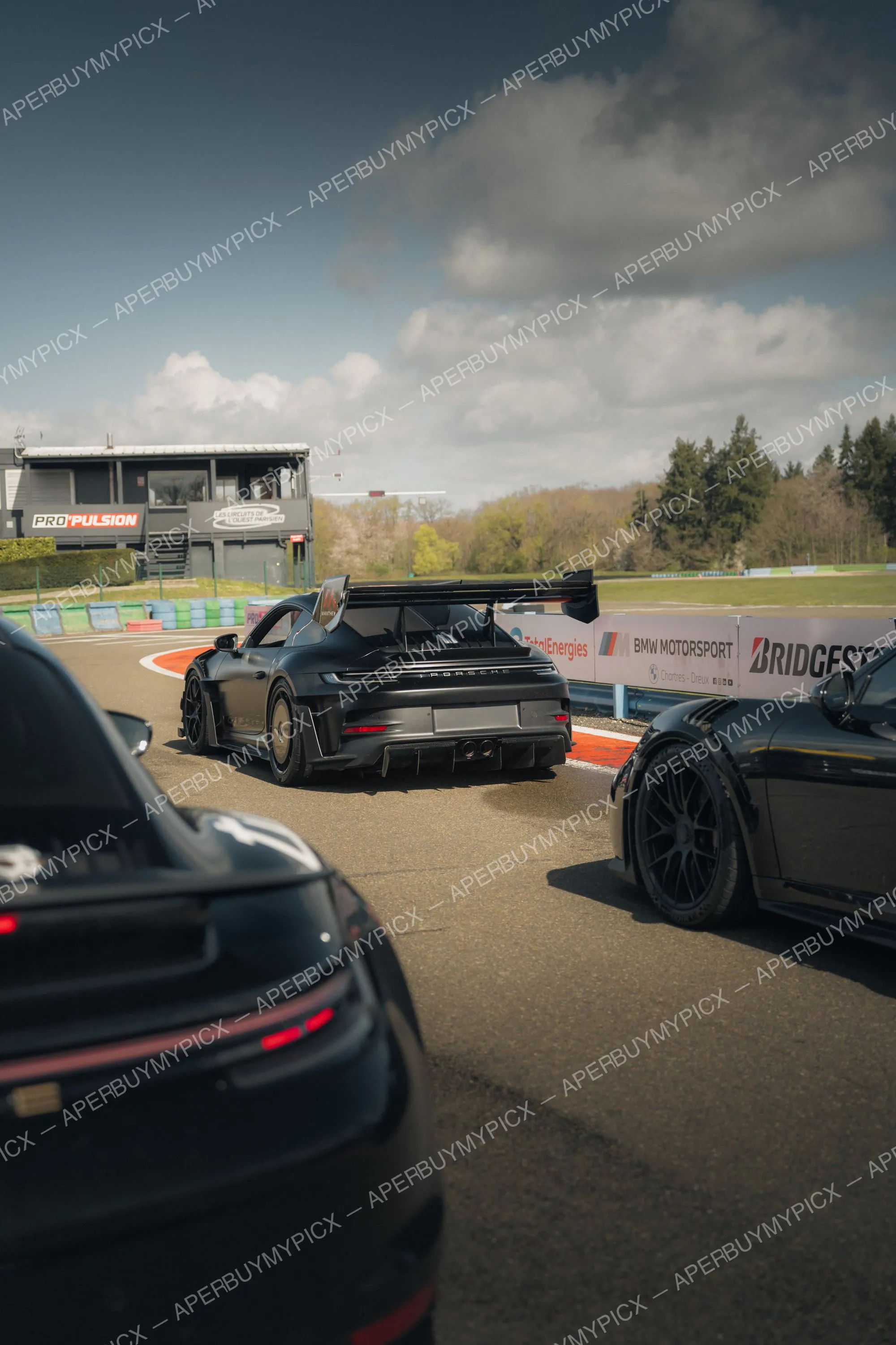 Porsche 911 GT3 noire avec aileron Manthey vue de l'arrière sur un circuit, entourée d'autres Porsche noires, panneaux BMW Motorsport et Bridgestone au fond sous un ciel nuageux.