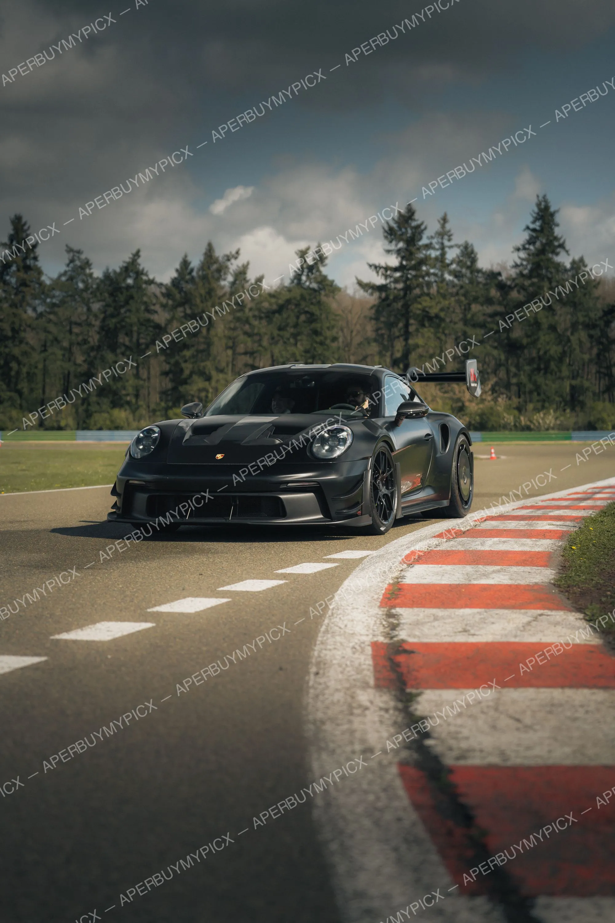 Porsche 911 GT3 RS noire mate roulant sur un circuit, sortie de virage sur les vibreurs rouges et blancs, forêt de sapins et ciel nuageux en arrière-plan