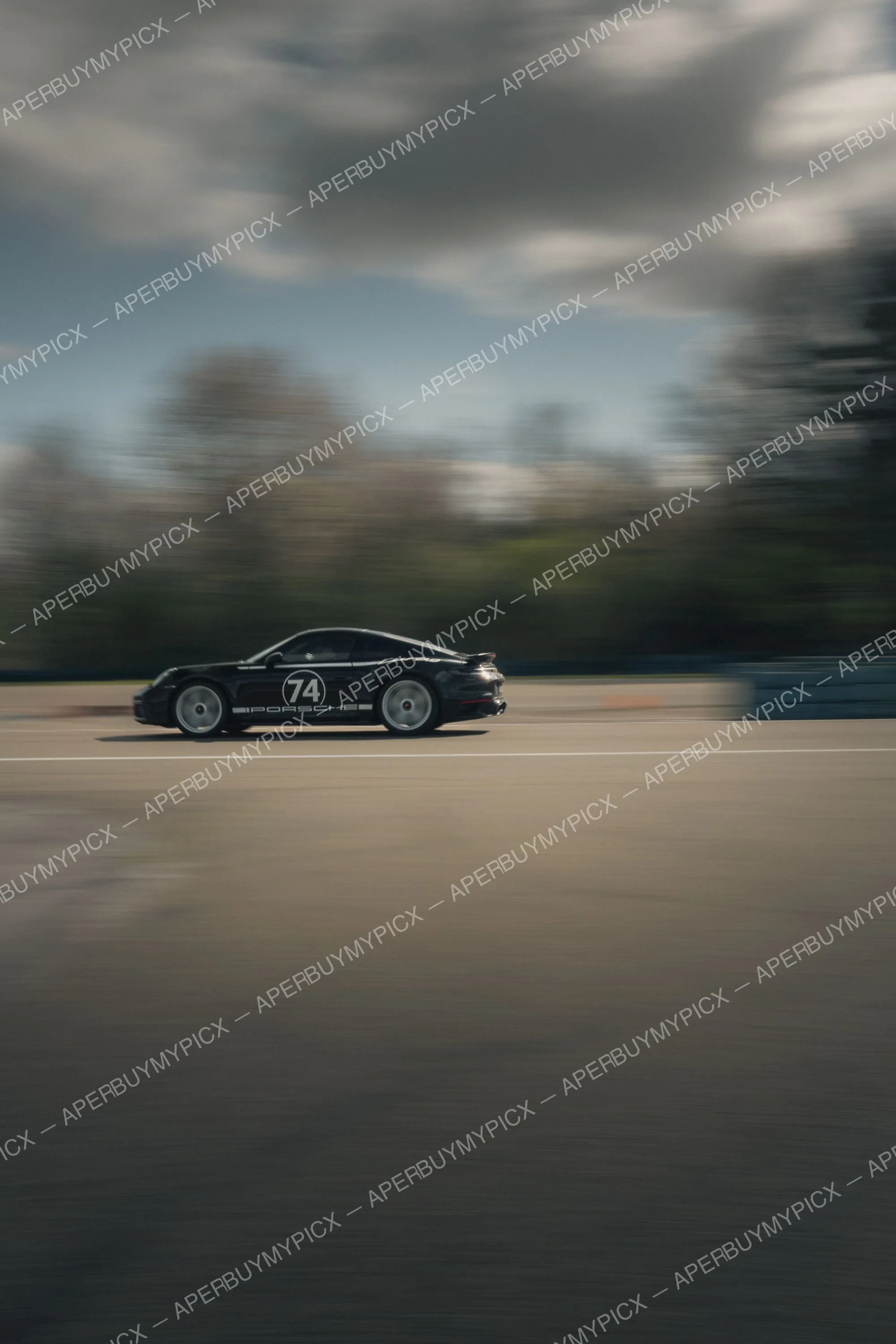 Porsche 911 noire portant le numéro 74 et le logo Porsche, roulant à grande vitesse sur un circuit avec un arrière-plan flou d'arbres et de ciel nuageux, effet de filé accentuant la vitesse.