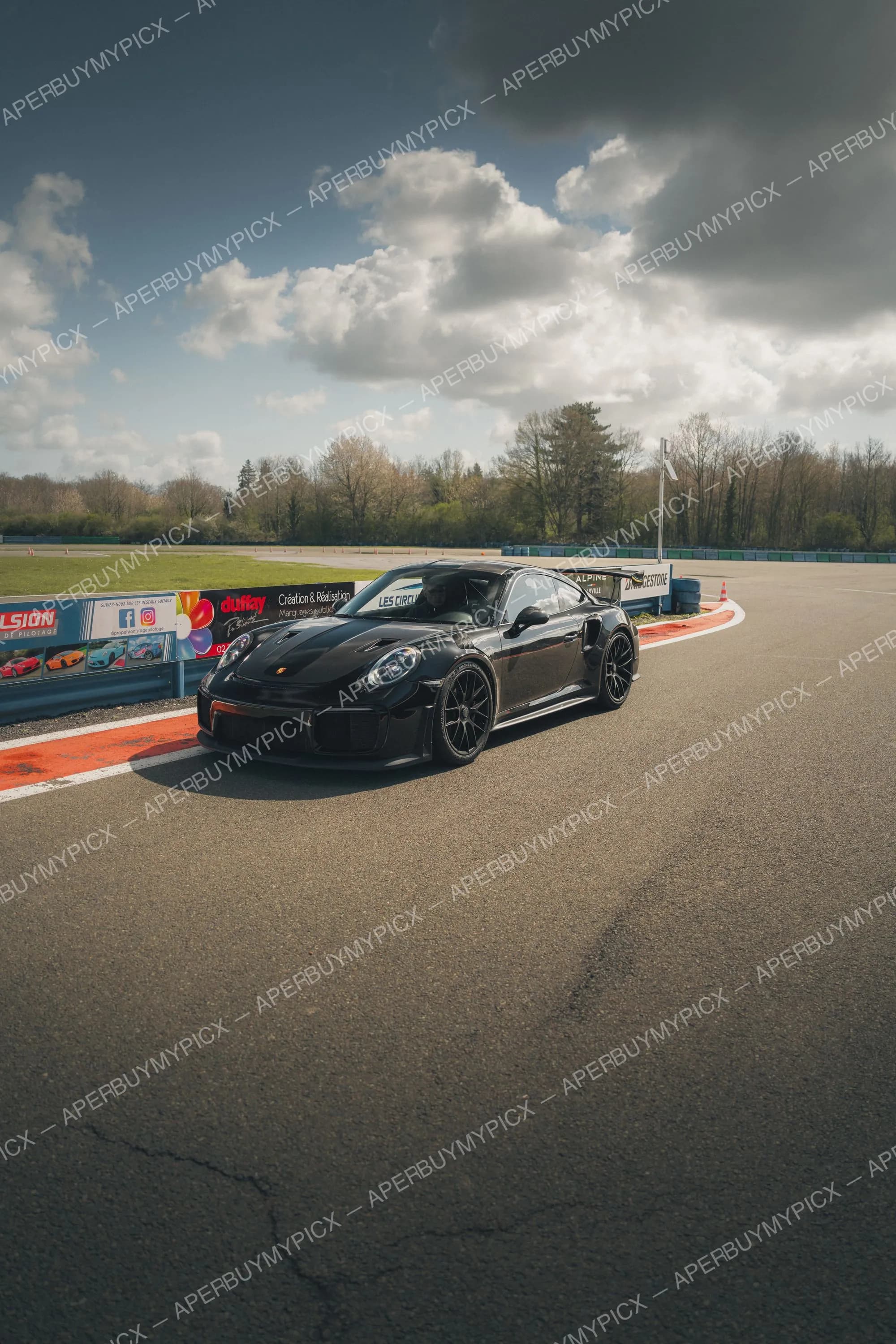 Porsche 911 GT2 RS noire à l'arrêt sur un circuit, jantes noires, aileron arrière, ciel nuageux et panneaux publicitaires en bordure de piste
