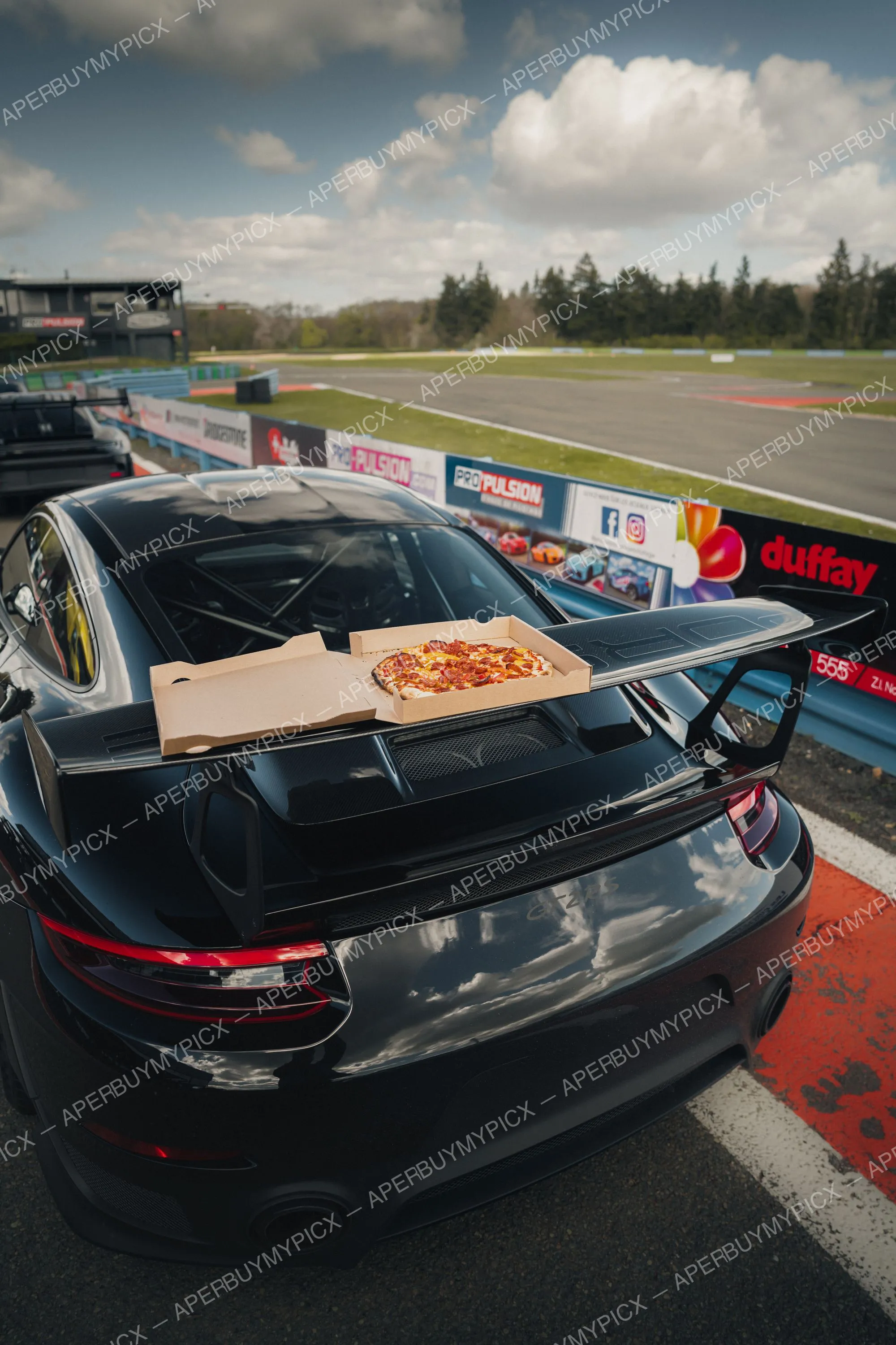 Porsche 911 GT2 RS noire vue arrière sur un circuit, avec une boîte de pizza pepperoni posée sur l'aileron en carbone, panneaux publicitaires et ciel nuageux en arrière-plan.
