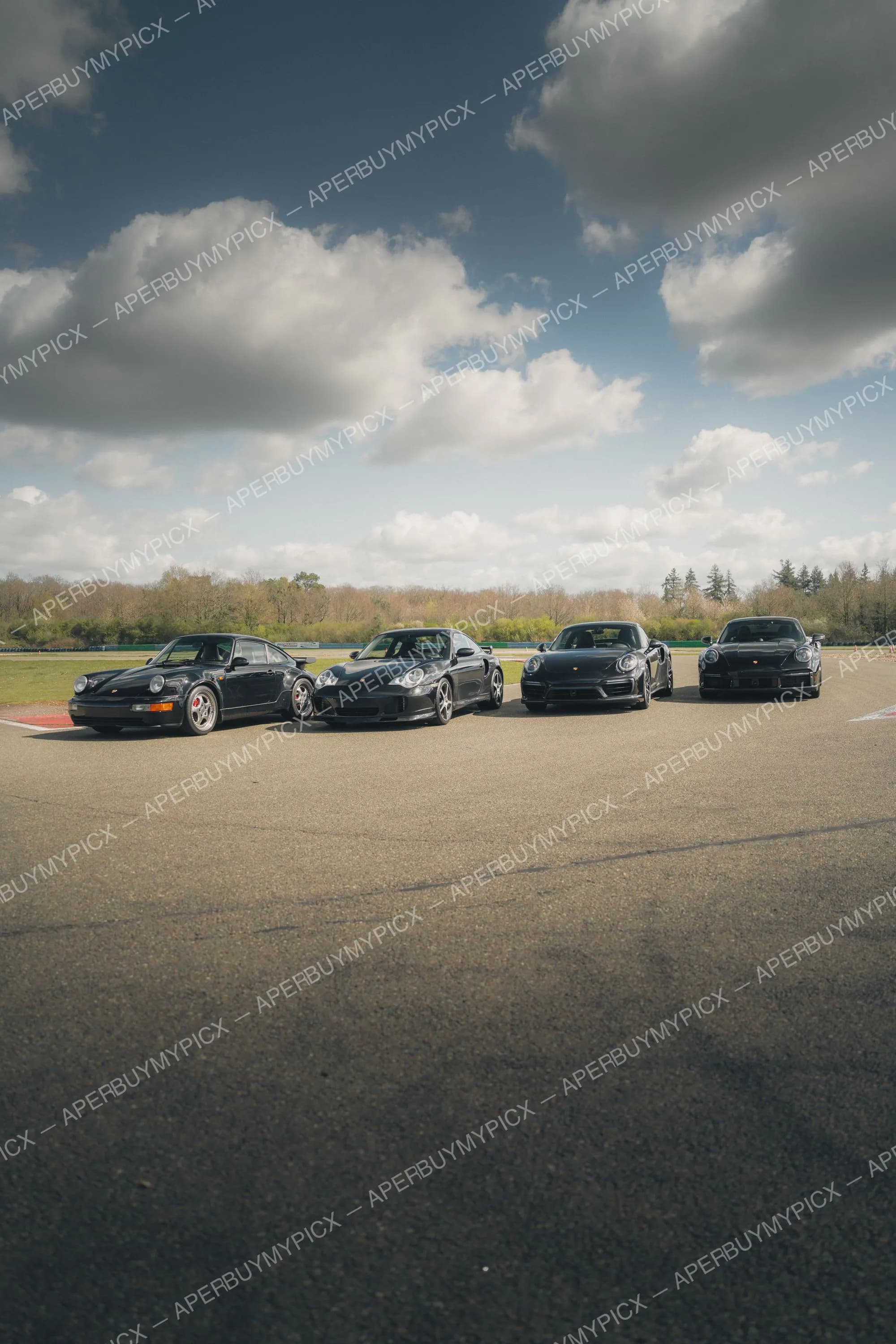 Quatre Porsche 911 Turbo noires de différentes générations alignées sur une piste de circuit sous un ciel nuageux, avec des arbres en arrière-plan.