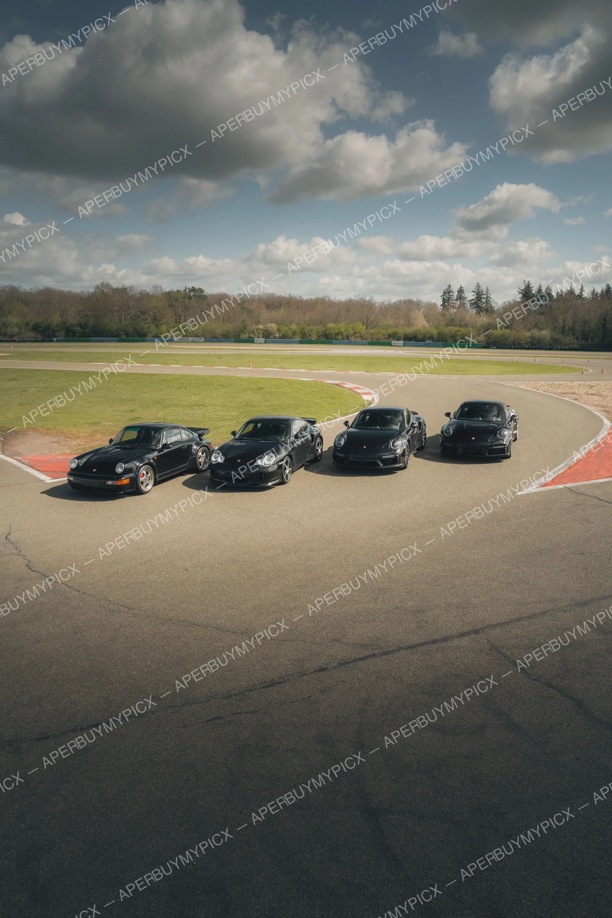 Quatre Porsche 911 Turbo noires de différentes générations alignées sur la piste d'un circuit, ciel nuageux et arbres en arrière-plan.