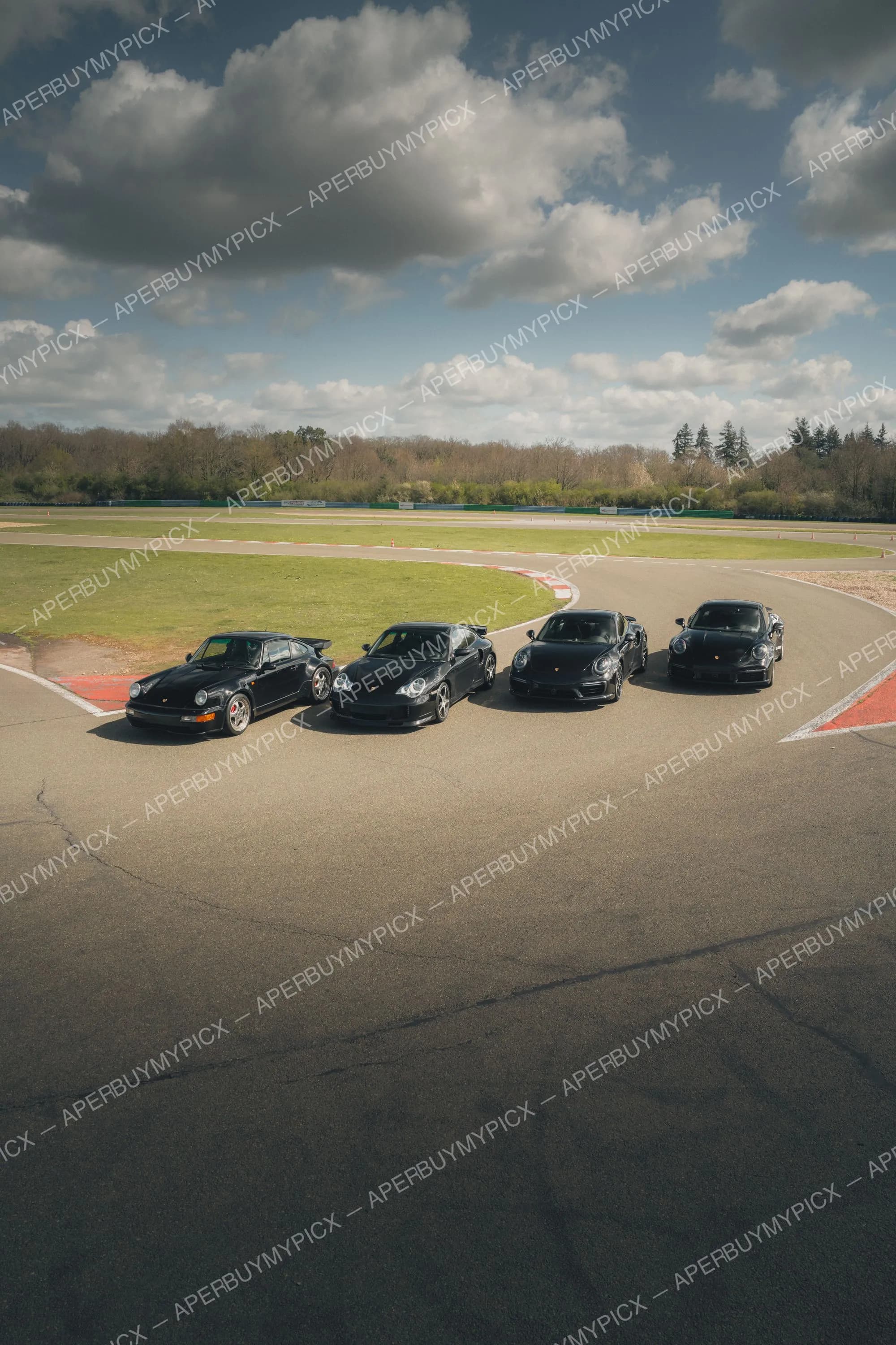 Quatre Porsche 911 noires de différentes générations alignées sur une piste de circuit sous un ciel nuageux, entourées de verdure.