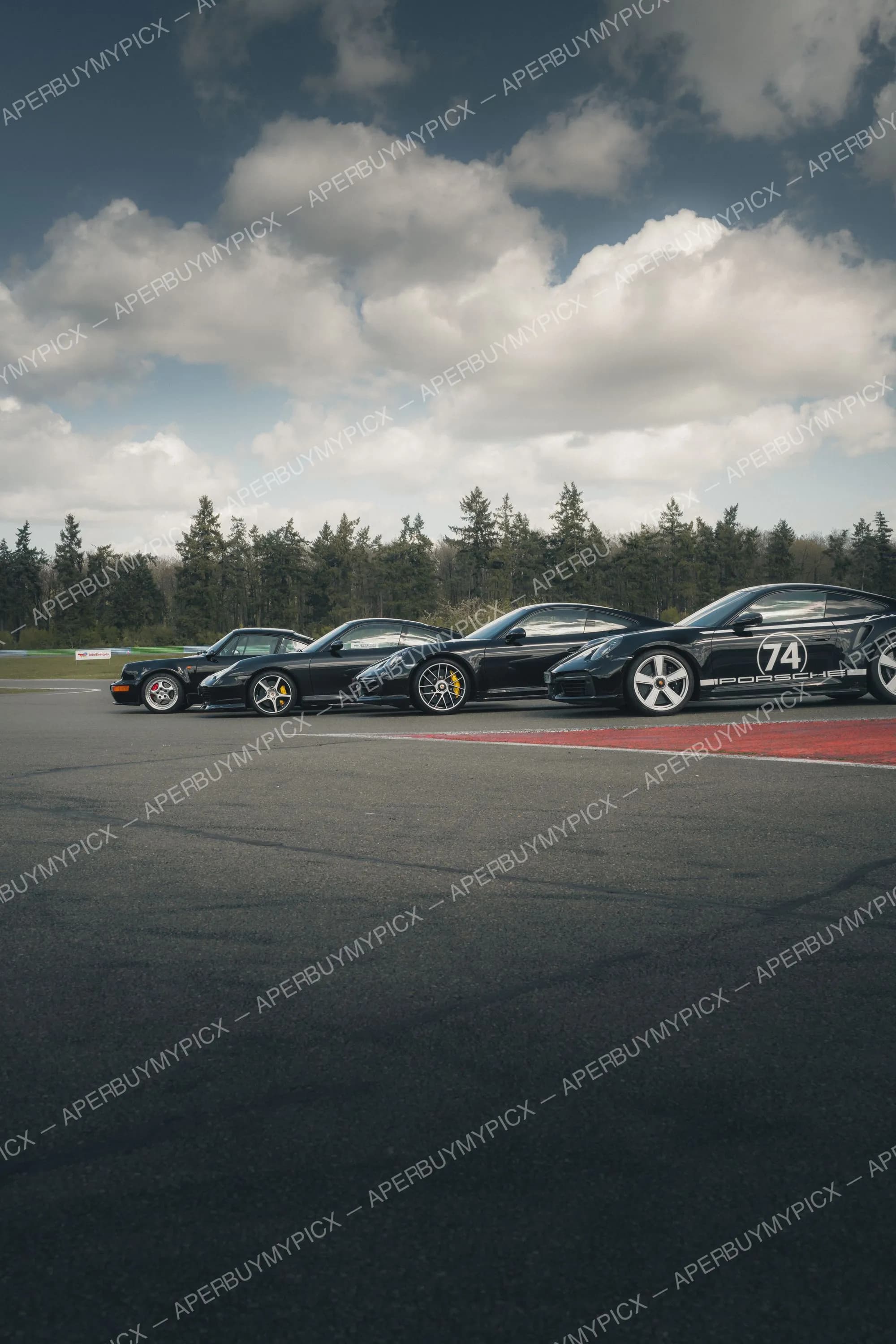 Quatre Porsche 911 Turbo noires, de la génération classique 993 à la moderne, alignées sur une piste de circuit sous un ciel nuageux, avec des arbres en arrière-plan.