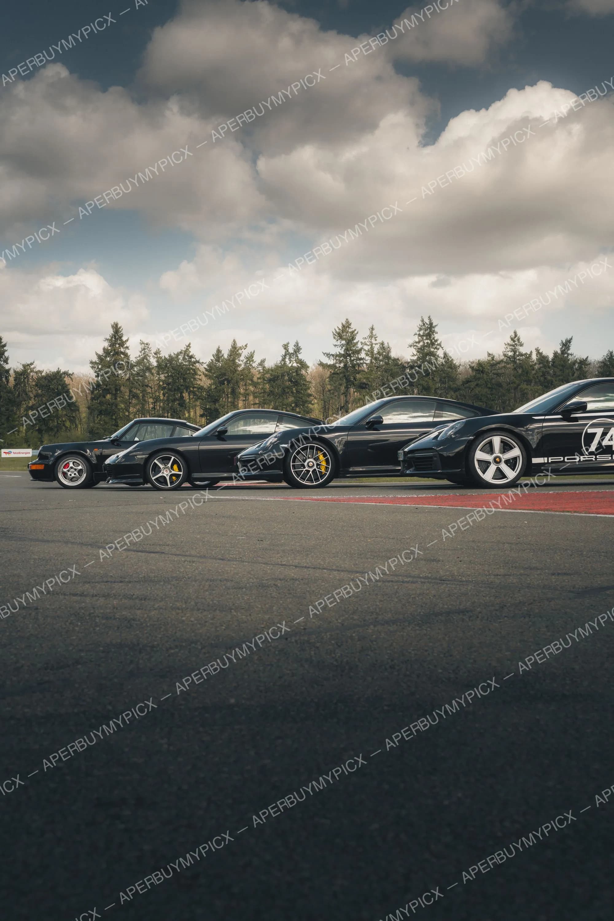 Quatre Porsche 911 noires de différentes générations alignées de profil sur un circuit sous un ciel nuageux, forêt en arrière-plan.