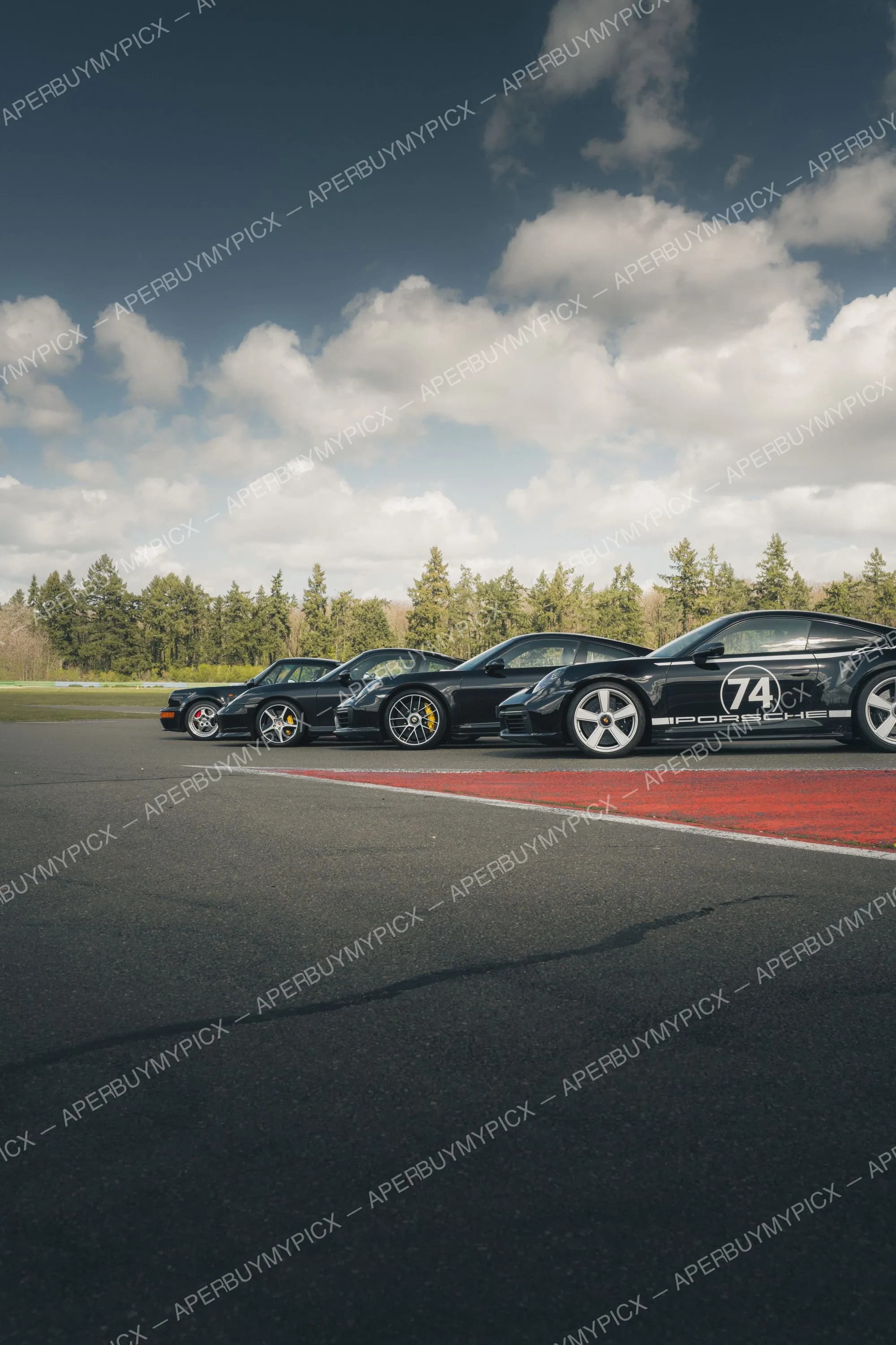 Quatre Porsche 911 noires de différentes générations alignées sur un circuit, une portant le numéro 74 et le logo Porsche, sous un ciel nuageux avec forêt en arrière-plan