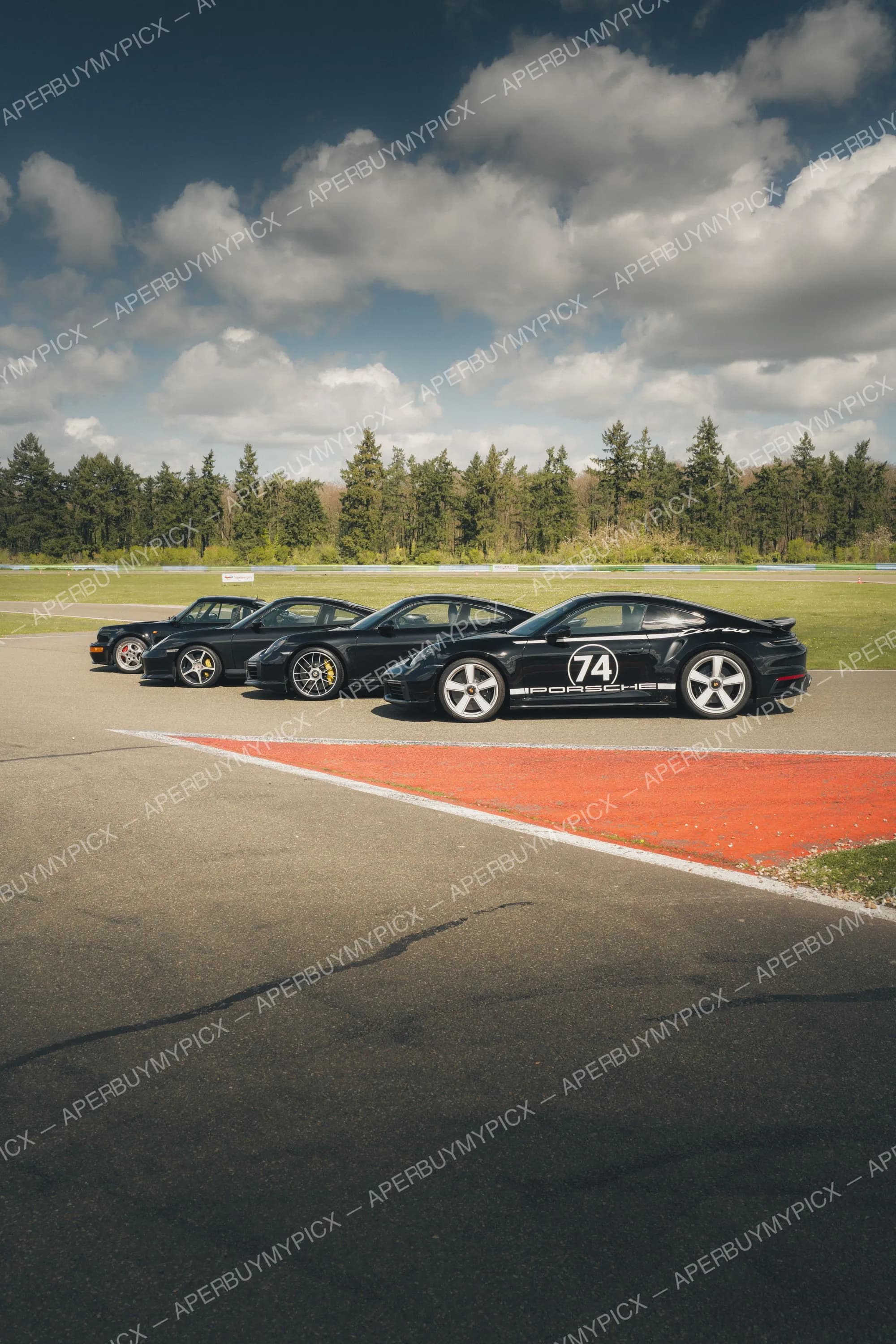 Quatre Porsche 911 Turbo noires de différentes générations alignées sur un circuit, la plus proche portant le numéro 74 et le logo Porsche, avec ciel nuageux et forêt en arrière-plan.