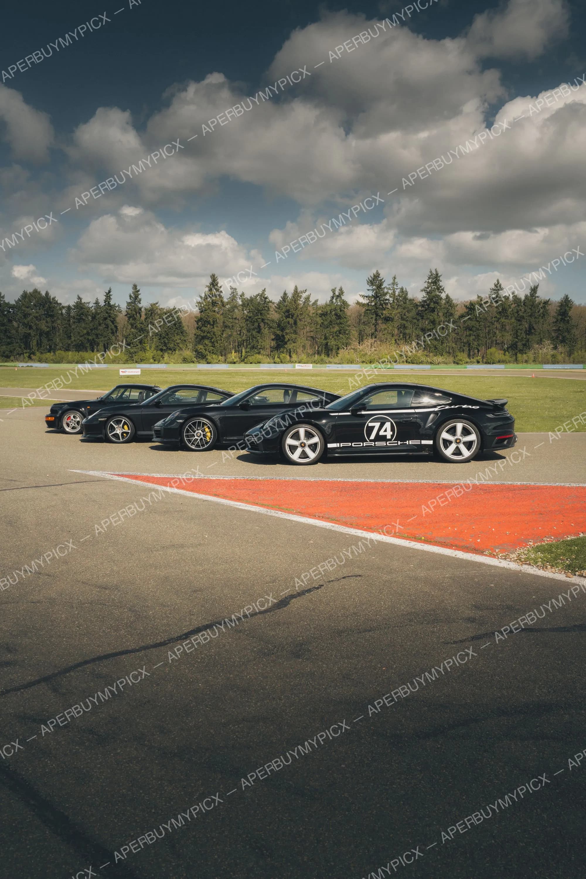 Quatre Porsche 911 Turbo noires de différentes générations alignées de profil sur un circuit, celle de devant portant le numéro 74 et le logo Porsche, sous un ciel nuageux avec forêt en arrière-plan.