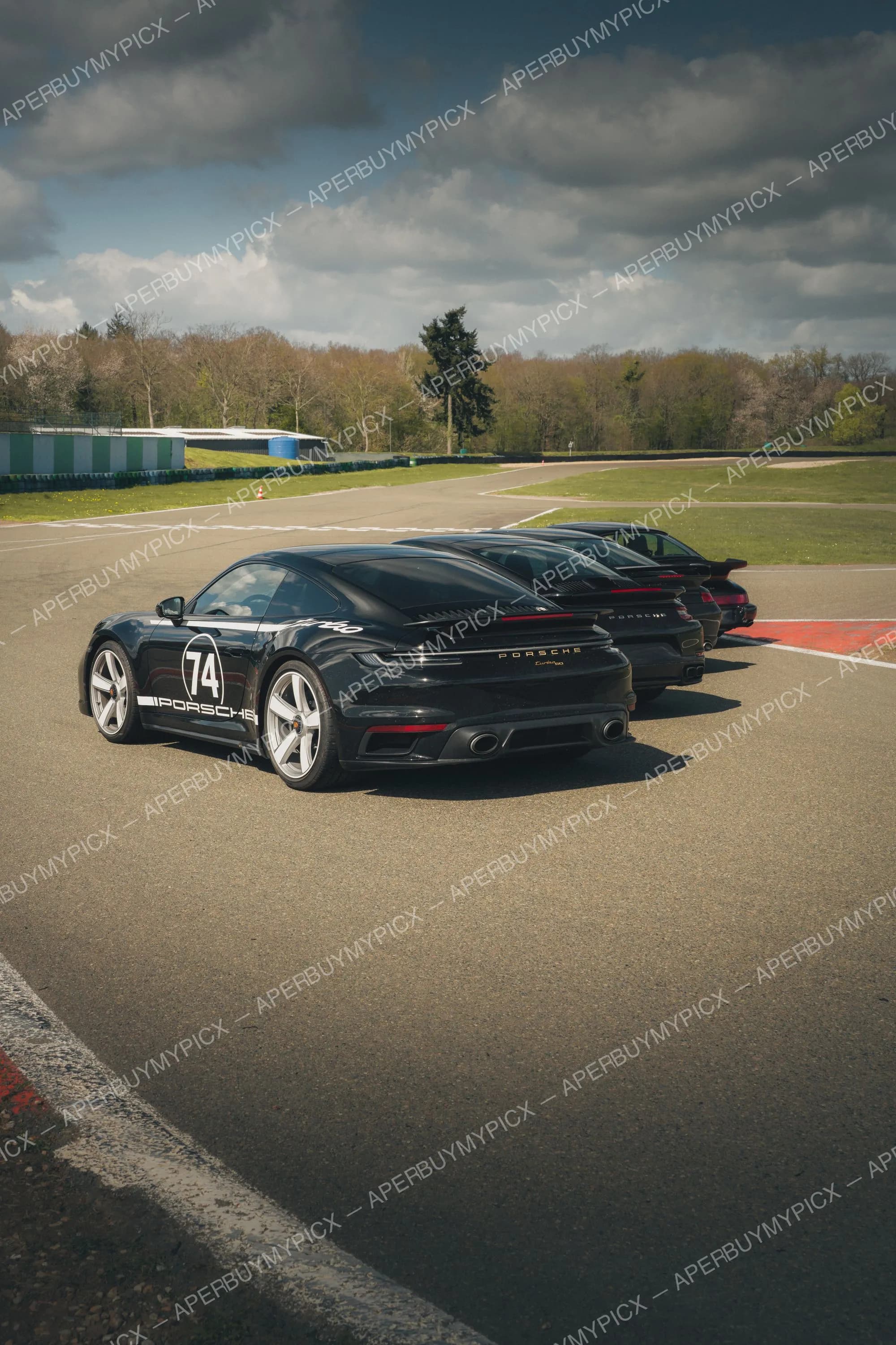 Trois Porsche 911 Turbo noires alignées sur une piste de circuit, la première portant le numéro 74 et la mention Porsche, sous un ciel nuageux avec arbres en arrière-plan.