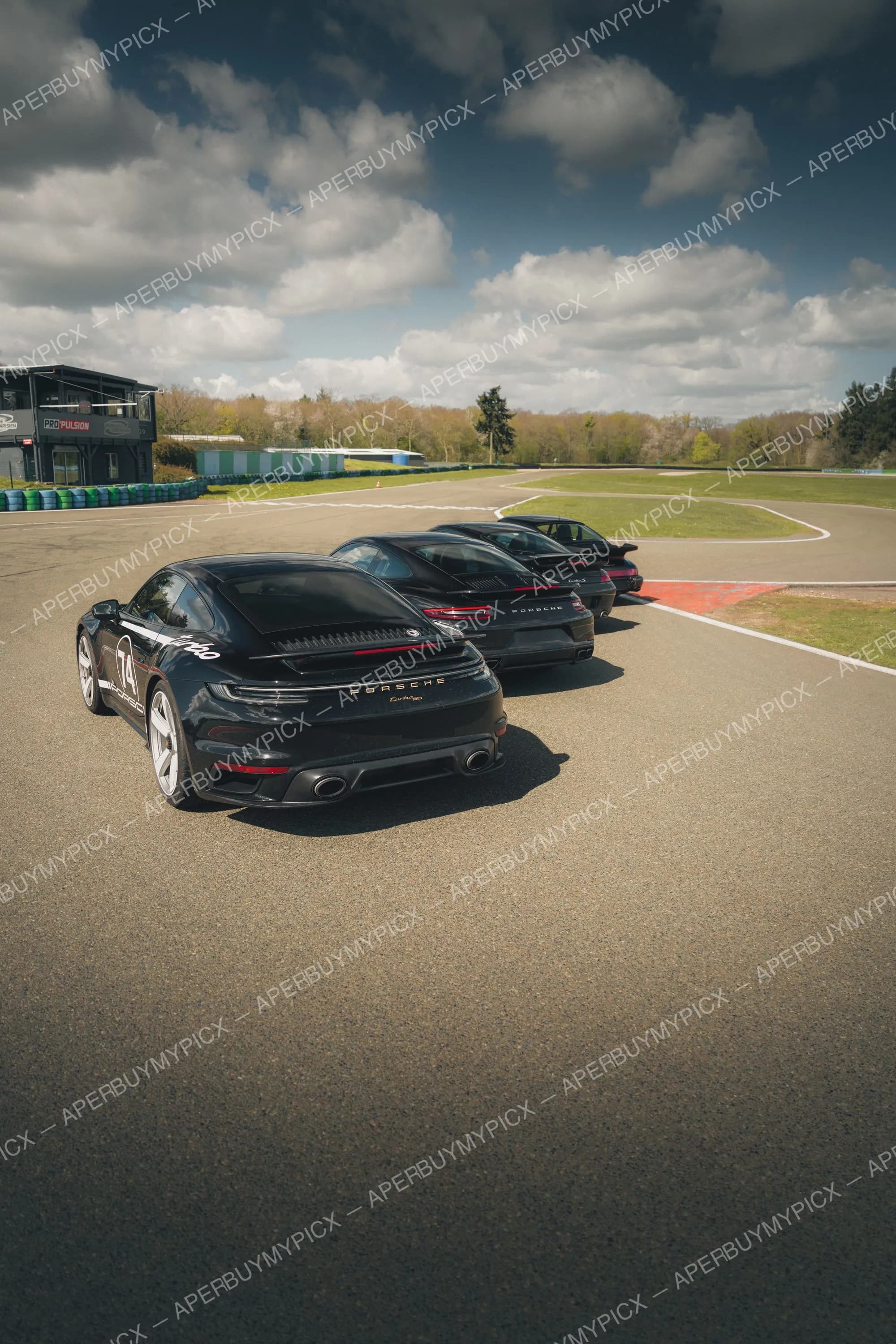 Trois Porsche 911 Turbo noires alignées sur une piste de circuit sous un ciel nuageux, vues de l'arrière, avec bâtiment et arbres en arrière-plan.