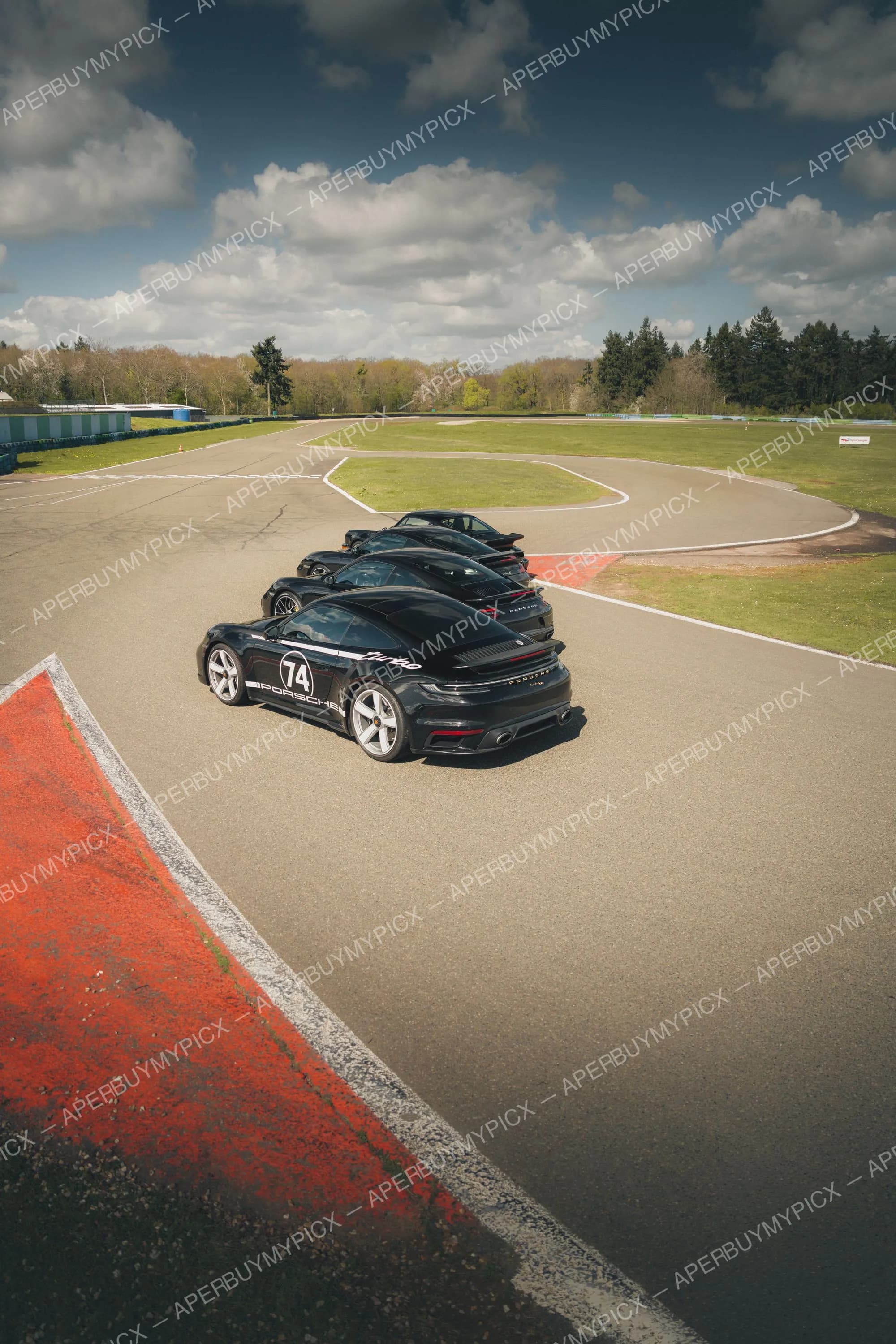 Trois Porsche 911 Turbo noires alignées sur un circuit, la première portant le numéro 74 et des logos Porsche, avec ciel nuageux et arbres en arrière-plan.