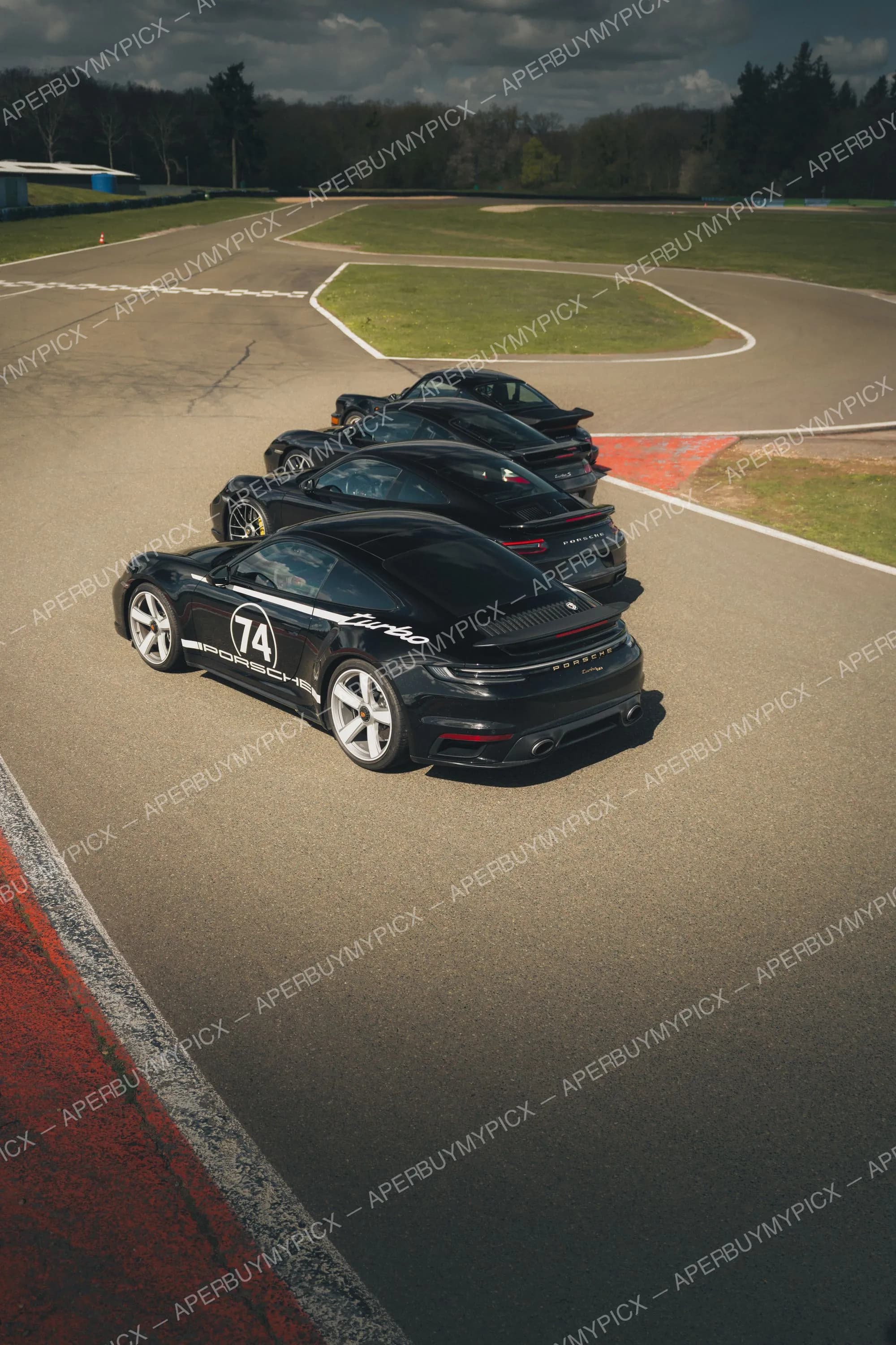 Quatre Porsche 911 Turbo noires alignées en file sur un circuit, la première portant le numéro 74 et un liseré blanc, sous un ciel nuageux avec forêt en arrière-plan