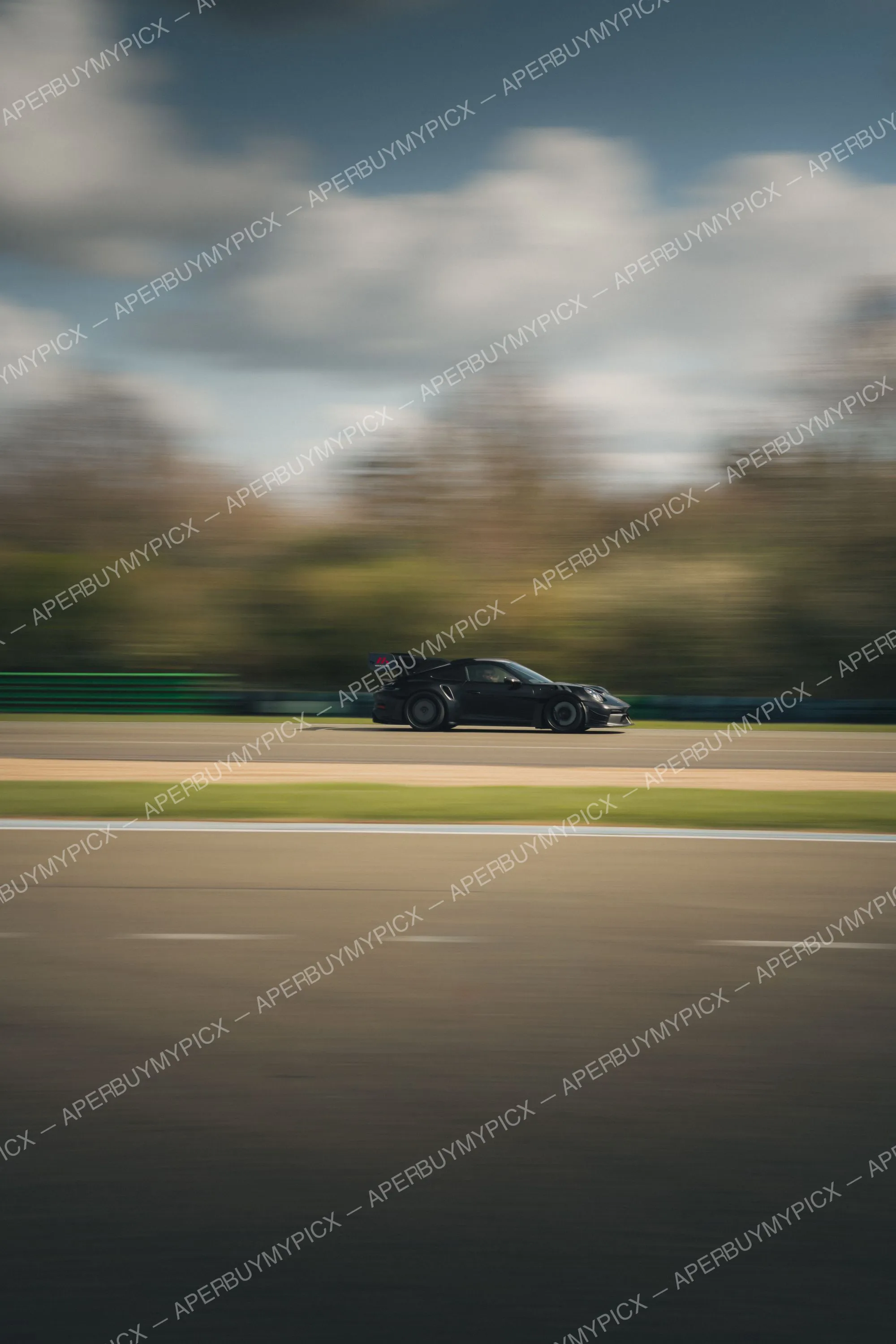 Porsche 911 GT3 RS noire avec aileron arrière roulant à grande vitesse sur un circuit, effet de filé dynamique avec arbres et ciel nuageux flous en arrière-plan