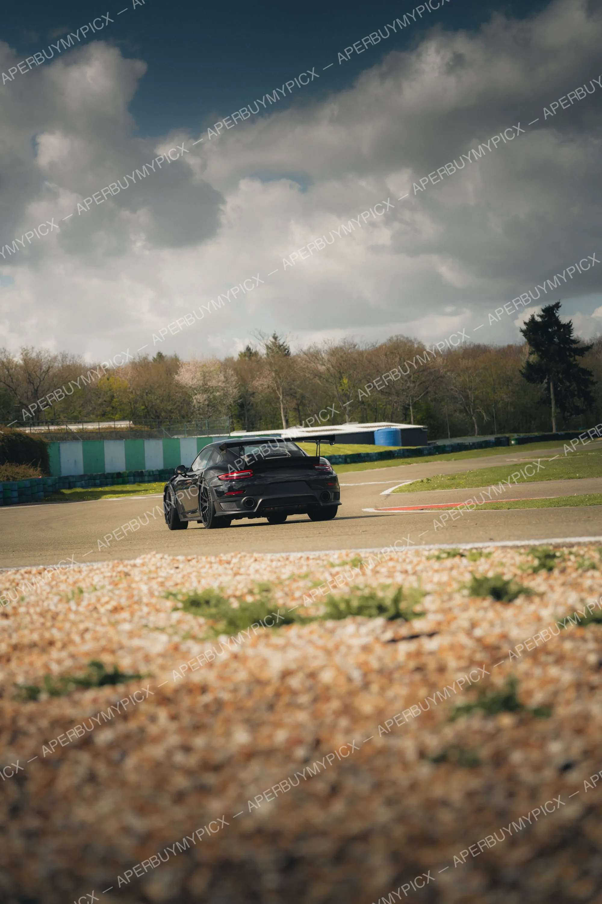 Porsche 911 GT2 RS noire avec aileron arrière roulant sur un circuit asphalté, vue arrière, sous un ciel nuageux avec arbres et bacs à gravier
