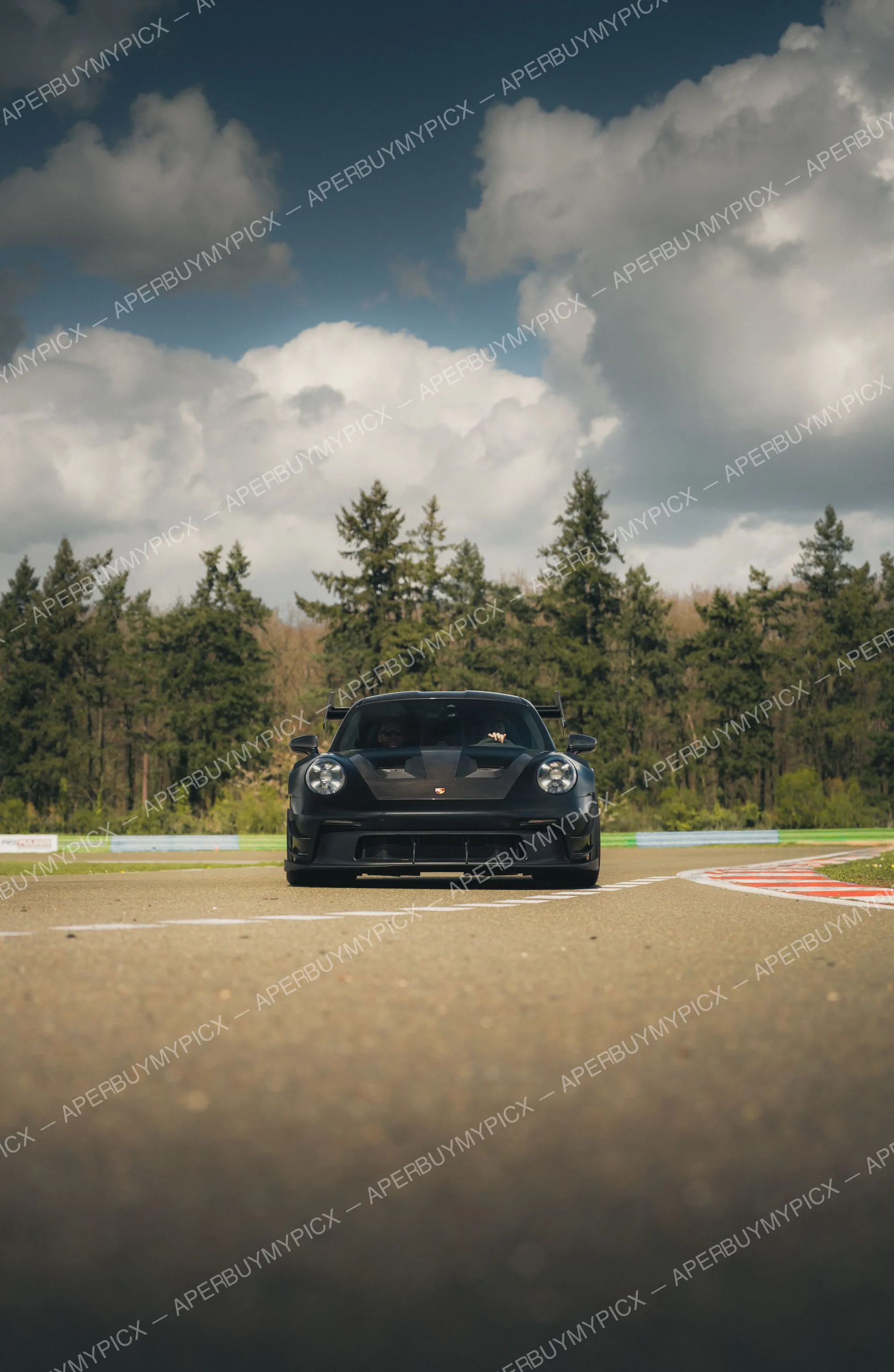 Porsche 911 GT3 RS noire vue de face sur une piste de circuit, avec vibreurs rouges et blancs, forêt de sapins et ciel nuageux en arrière-plan