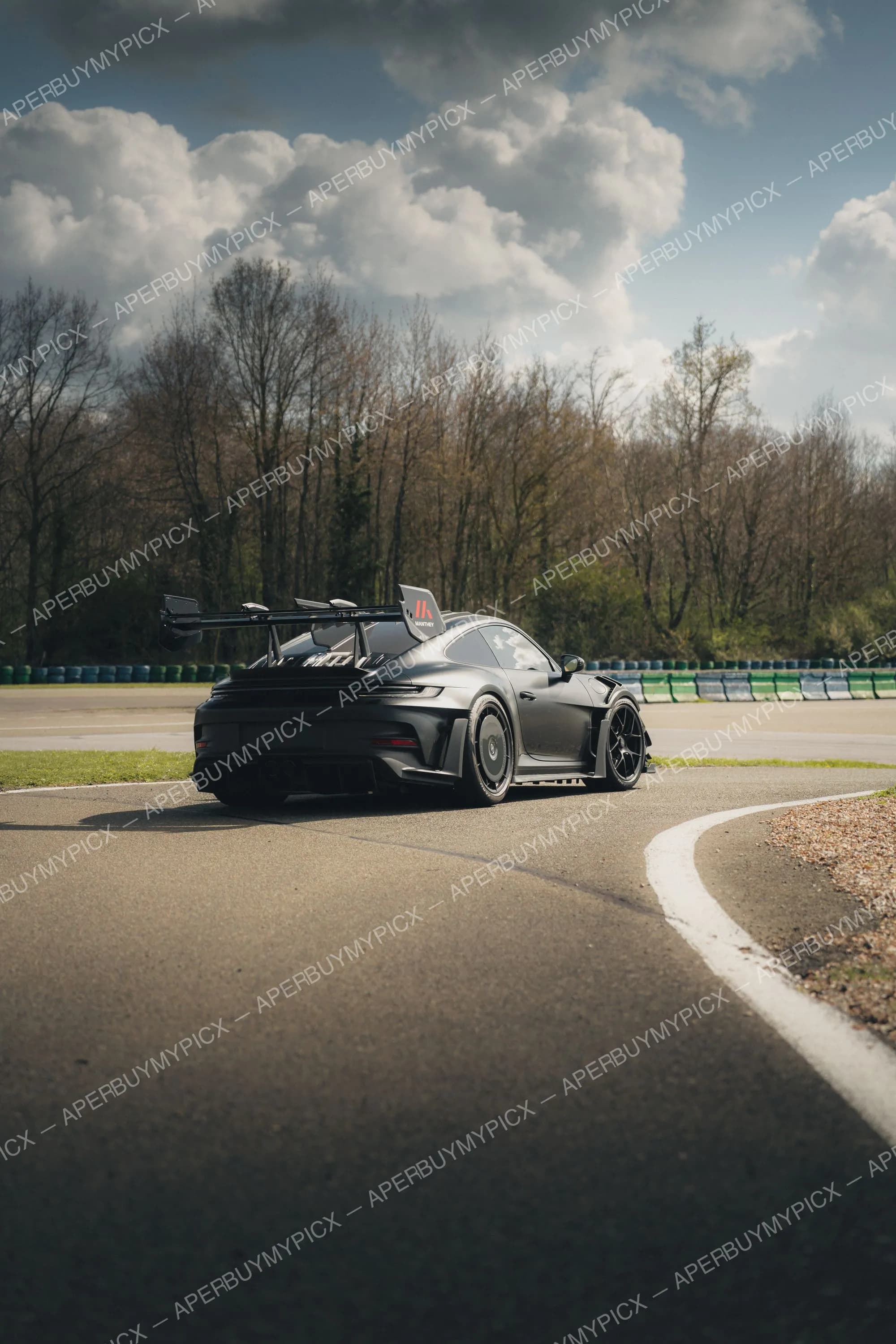 Porsche 911 GT3 RS noire équipée du kit Manthey avec aileron arrière, vue de trois quarts arrière sur un circuit bordé d'arbres sous un ciel nuageux