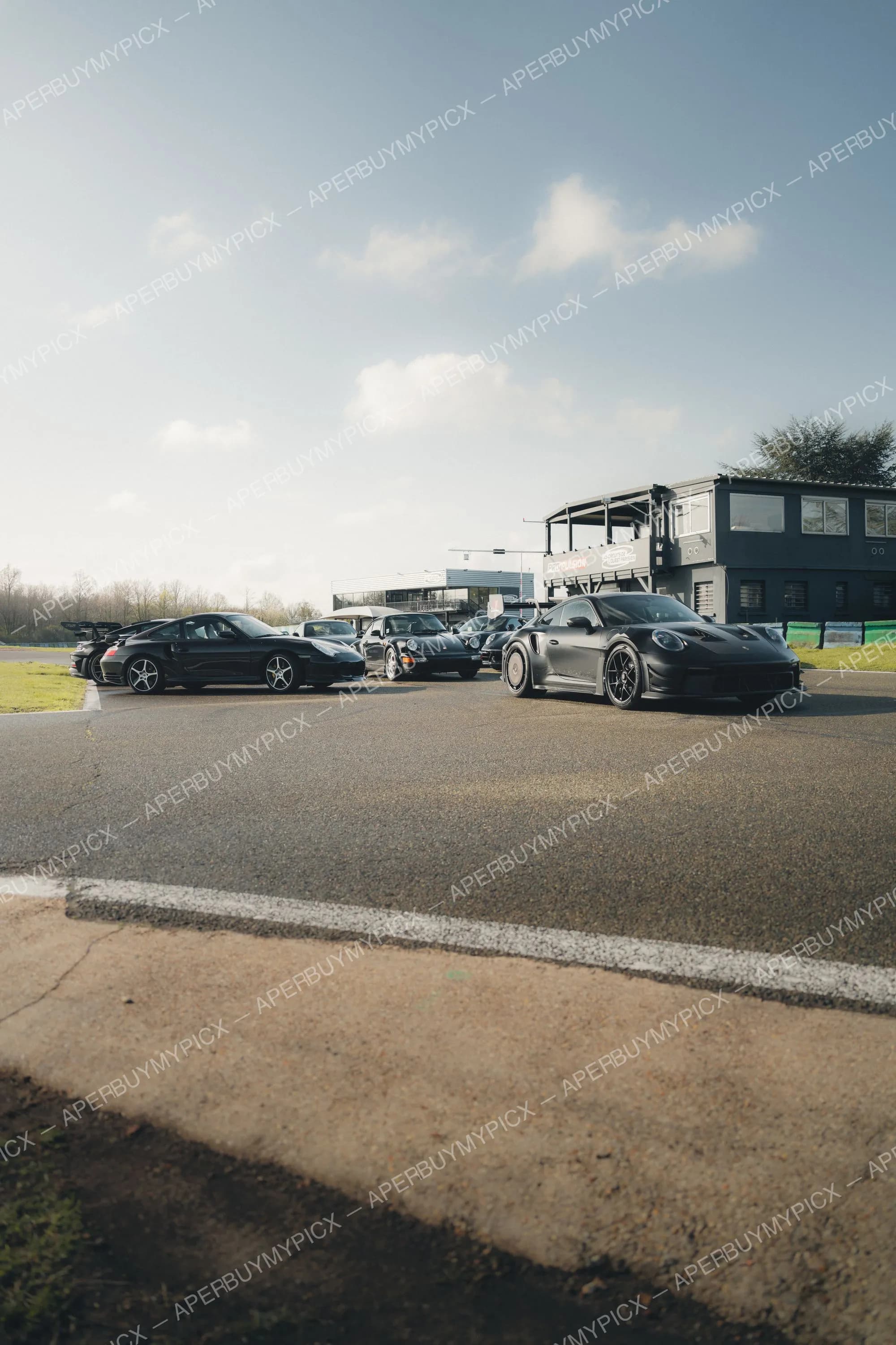 Plusieurs Porsche 911 noires alignées sur la piste d'un circuit automobile, dont une 911 Turbo mate au premier plan, avec des bâtiments de paddock et un ciel nuageux en arrière-plan.