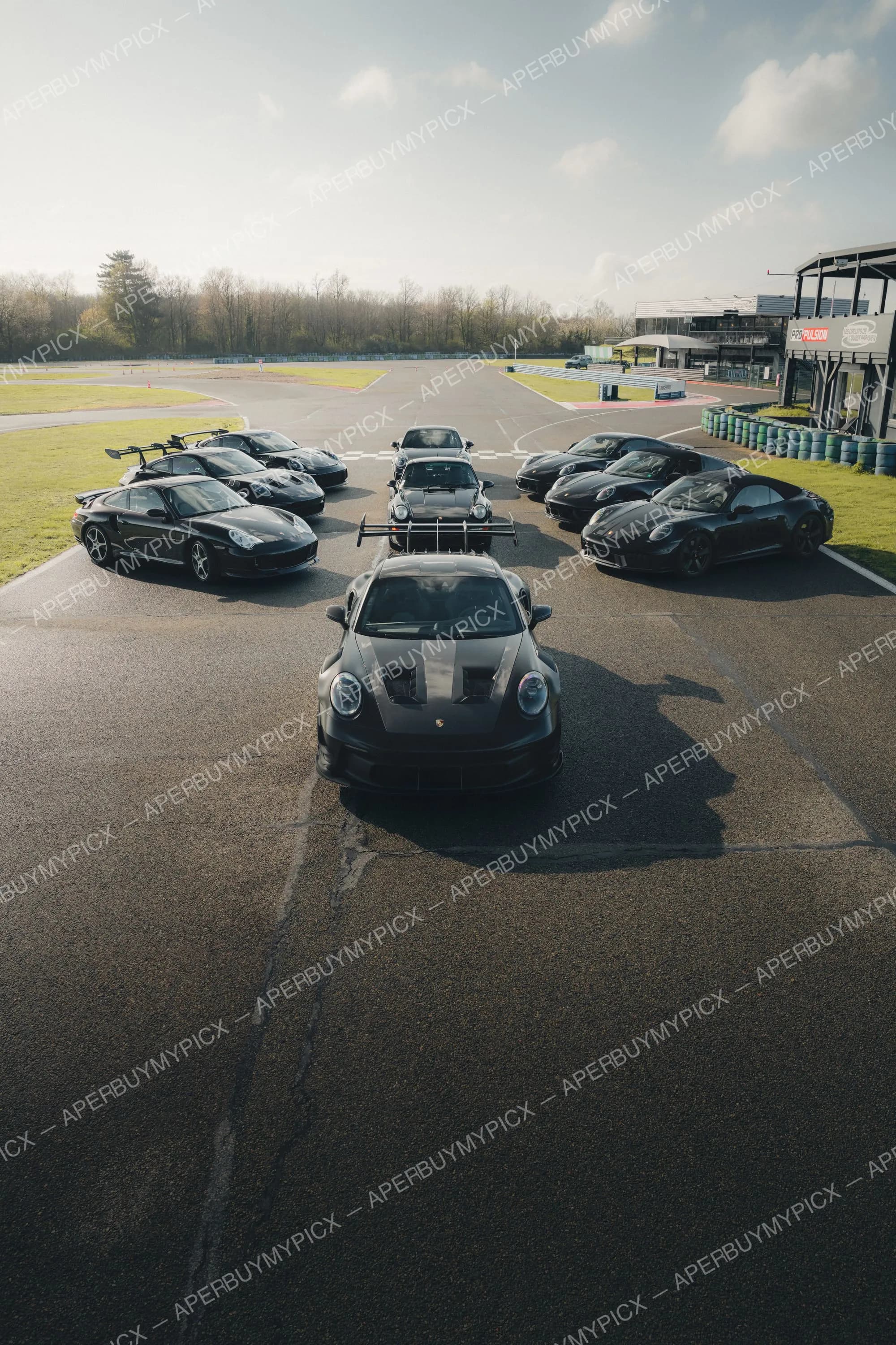 Rassemblement de plusieurs Porsche 911 noires, dont une GT3 RS en formation avec aileron, disposées en V sur la piste d'un circuit automobile avec les bâtiments et pneus en arrière-plan sous un ciel nuageux.