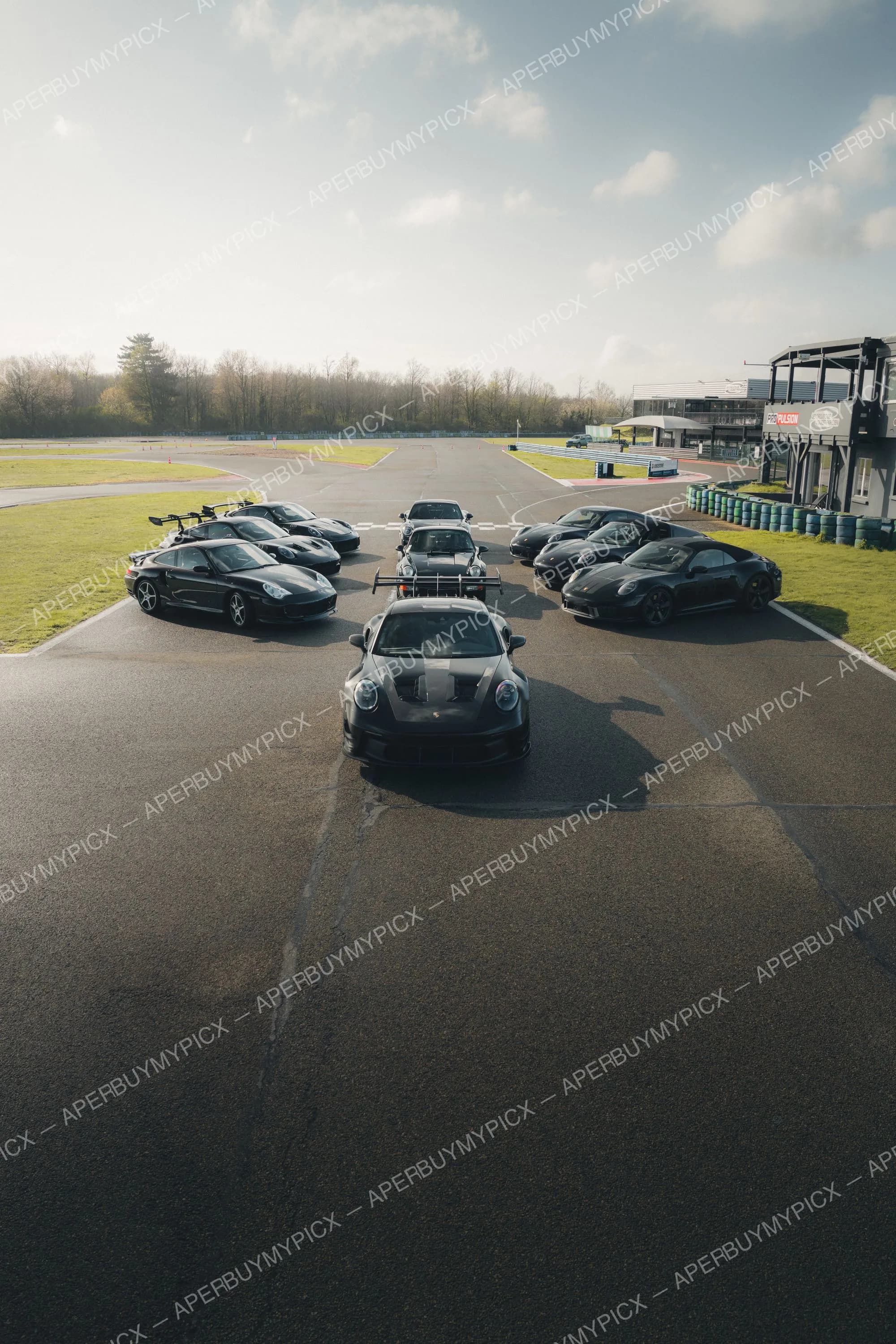 Groupe de voitures Porsche 911 noires disposées en formation sur la piste d'un circuit automobile, sous un ciel nuageux, avec un modèle GT3 RS à aileron au premier plan et des bâtiments de paddock à droite.