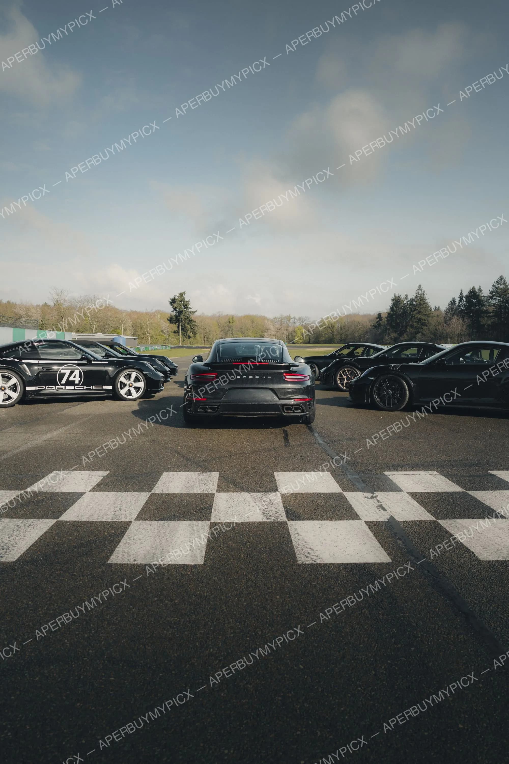 Groupe de Porsche 911 noires garées sur un circuit de course derrière une ligne d'arrivée à damier, sous un ciel nuageux avec forêt en arrière-plan