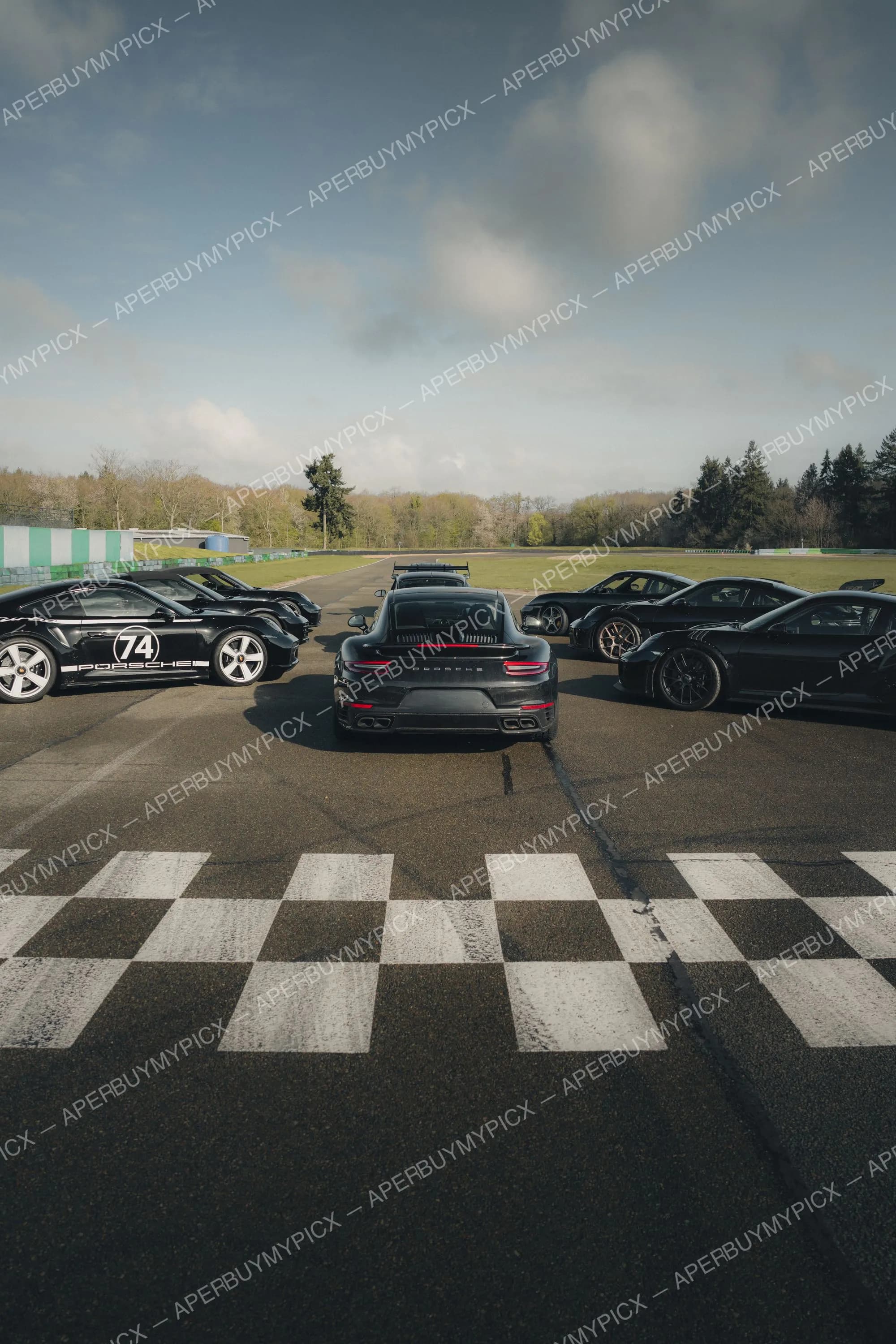 Groupe de Porsche 911 noires alignées sur un circuit automobile, vue depuis la ligne d'arrivée à damier, avec forêt et ciel nuageux en arrière-plan