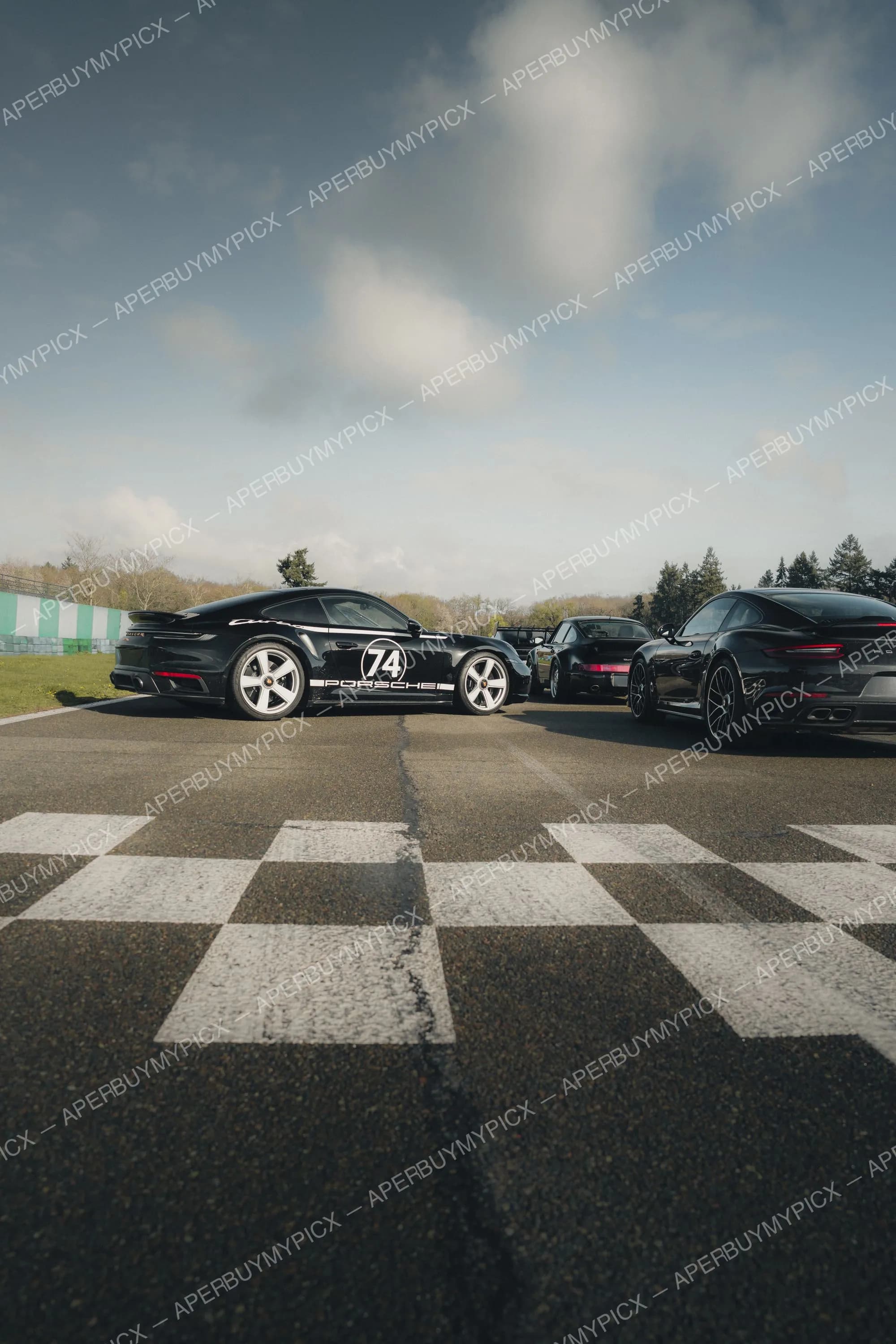 Trois Porsche 911 noires stationnées sur une piste de course derrière une ligne d'arrivée à damier, dont une avec le numéro 74 et le logo Porsche, sous un ciel nuageux avec arbres en arrière-plan.