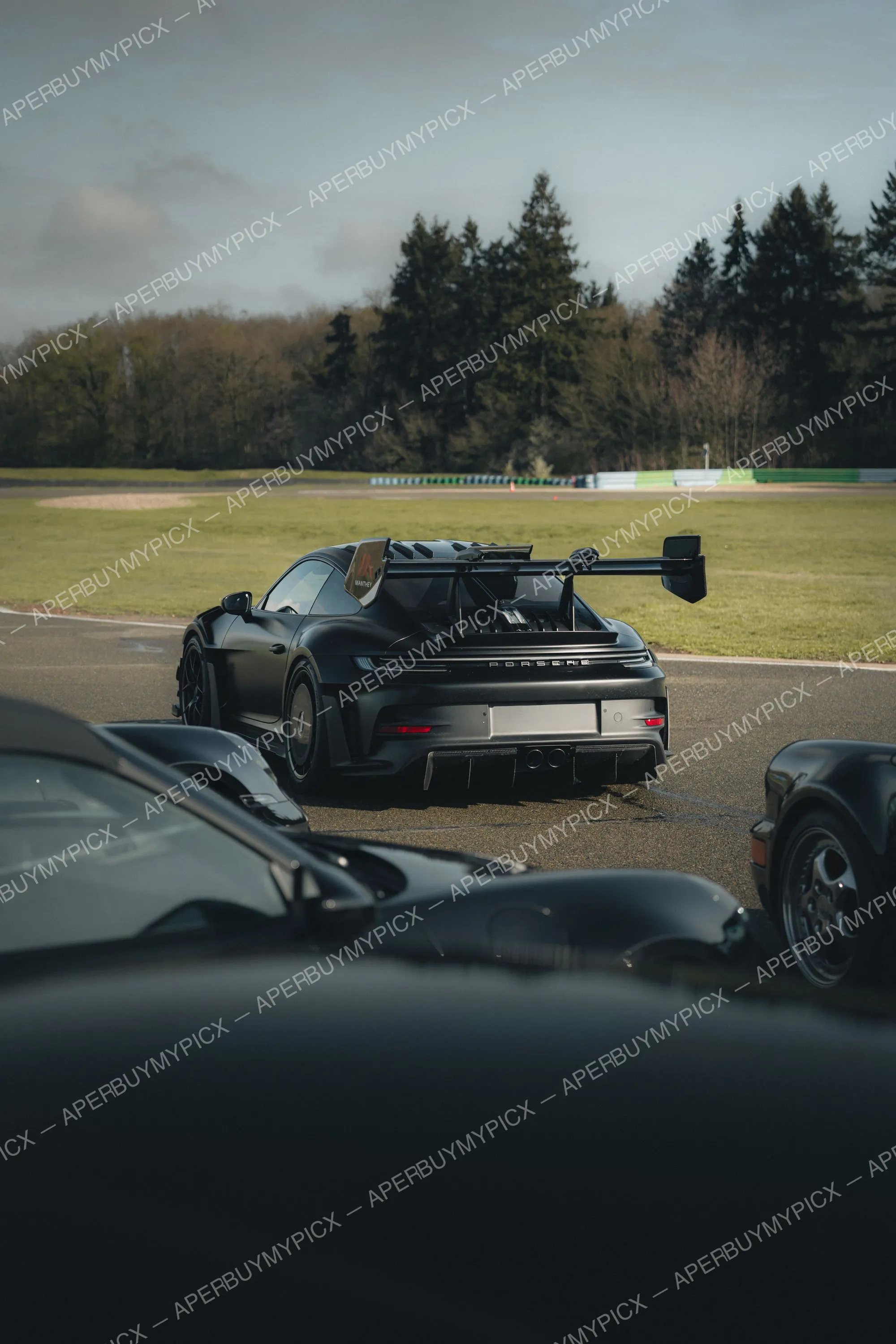 Porsche 911 GT3 RS noire équipée du kit Manthey vue de l'arrière sur un circuit, aileron arrière imposant, garée sur la piste devant une pelouse et une forêt de sapins sous un ciel nuageux.