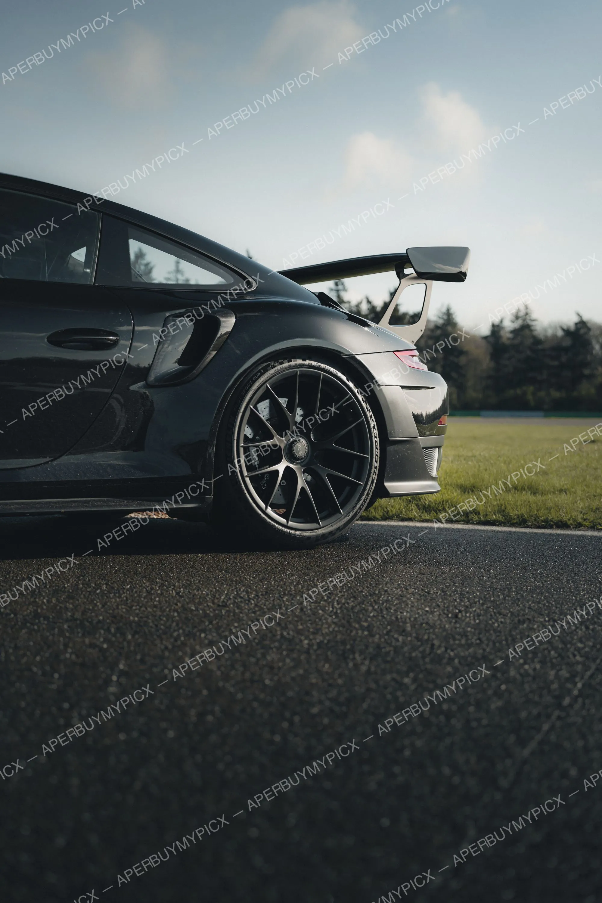 Porsche 911 GT3 RS noire vue de profil arrière avec aileron et jantes noires, stationnée sur un circuit au coucher du soleil