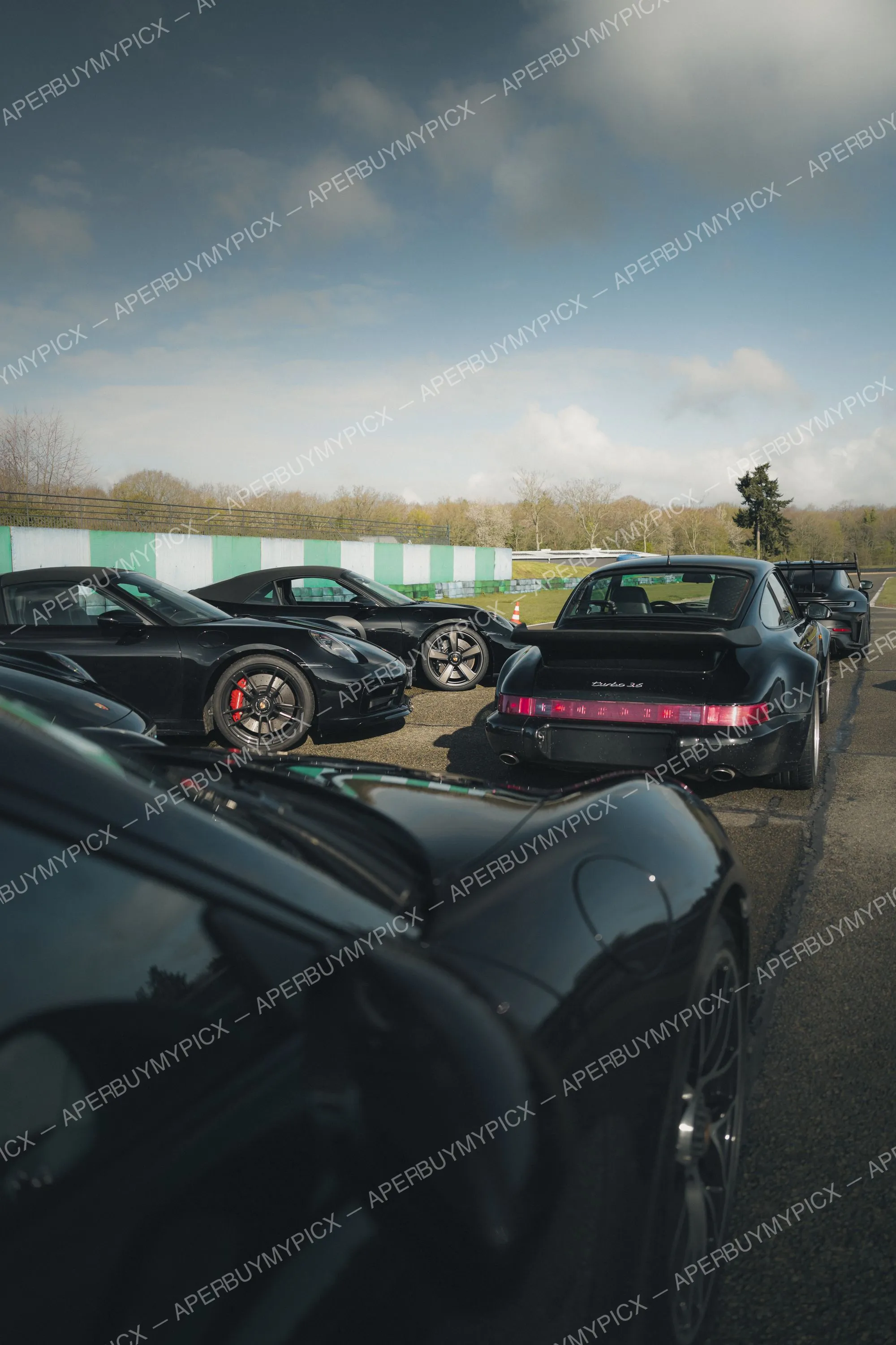 Plusieurs Porsche 911 noires, dont une 911 Turbo 3.6 classique avec aileron, garées sur un circuit sous un ciel nuageux, mur vert et blanc en arrière-plan.