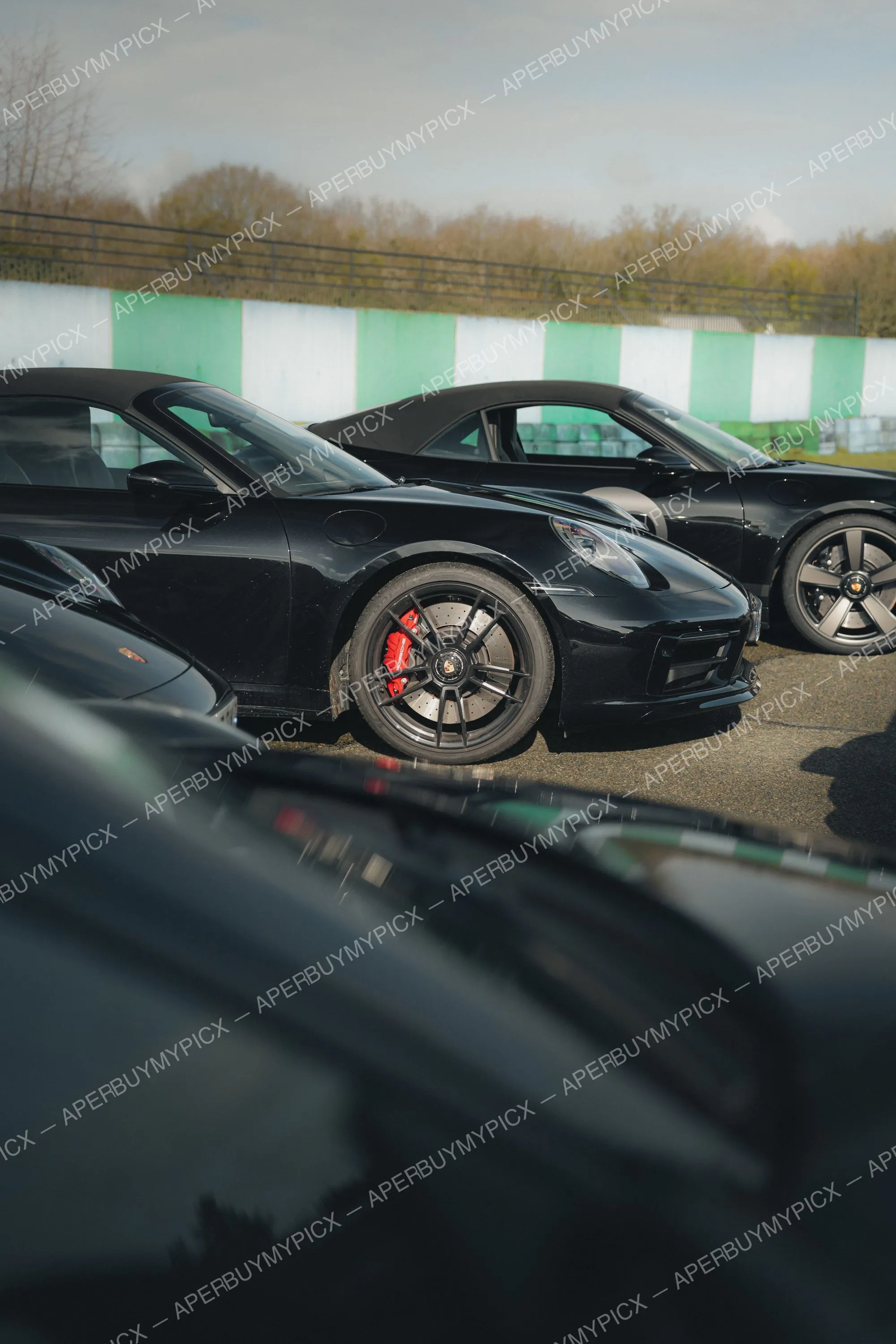 Porsche 911 cabriolet noire avec étriers de frein rouges et jantes noires, garée sur un circuit devant un muret vert et blanc.