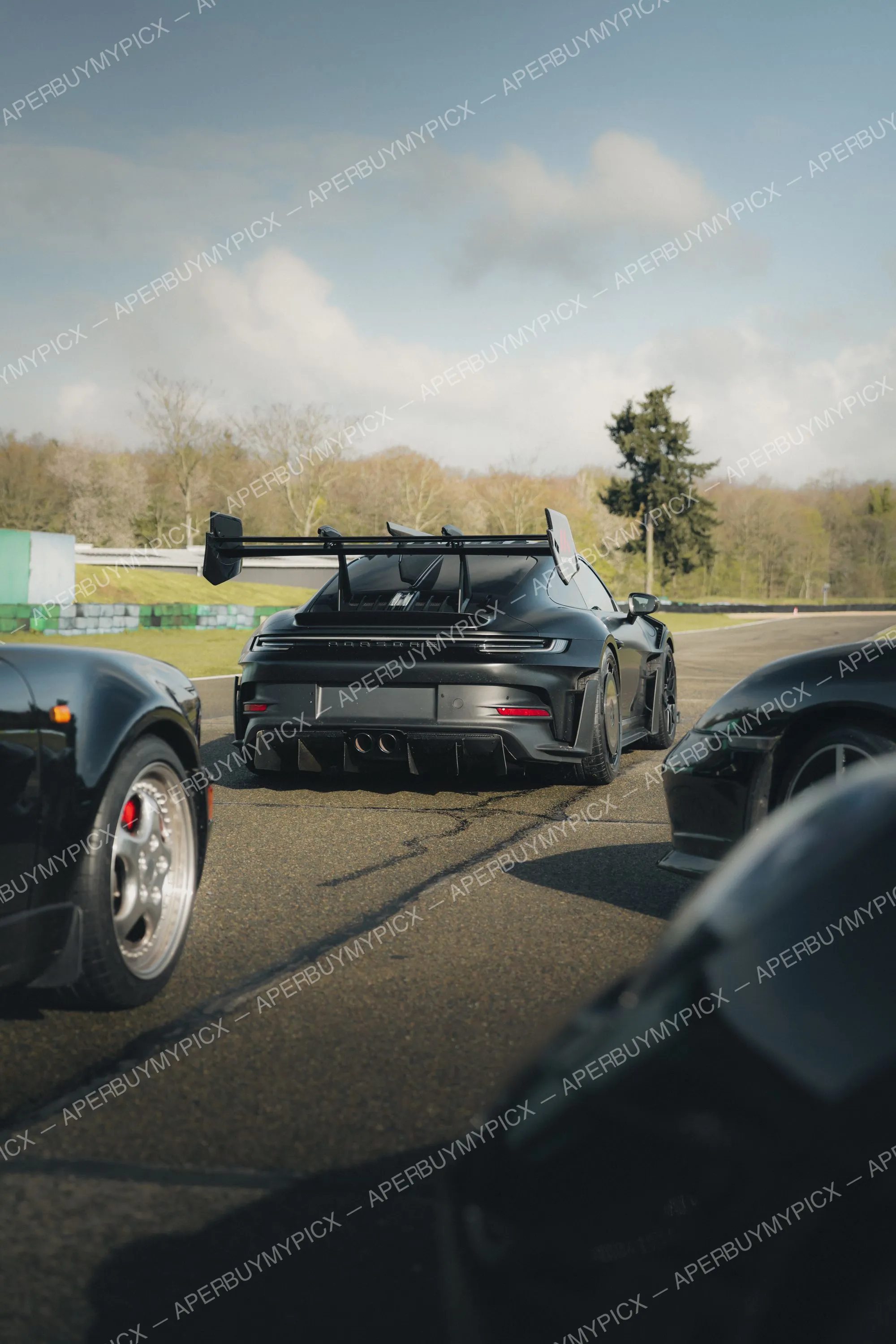 Porsche 911 GT3 RS noire vue de l'arrière avec aileron sur un circuit, entourée de deux autres voitures de sport noires par temps nuageux