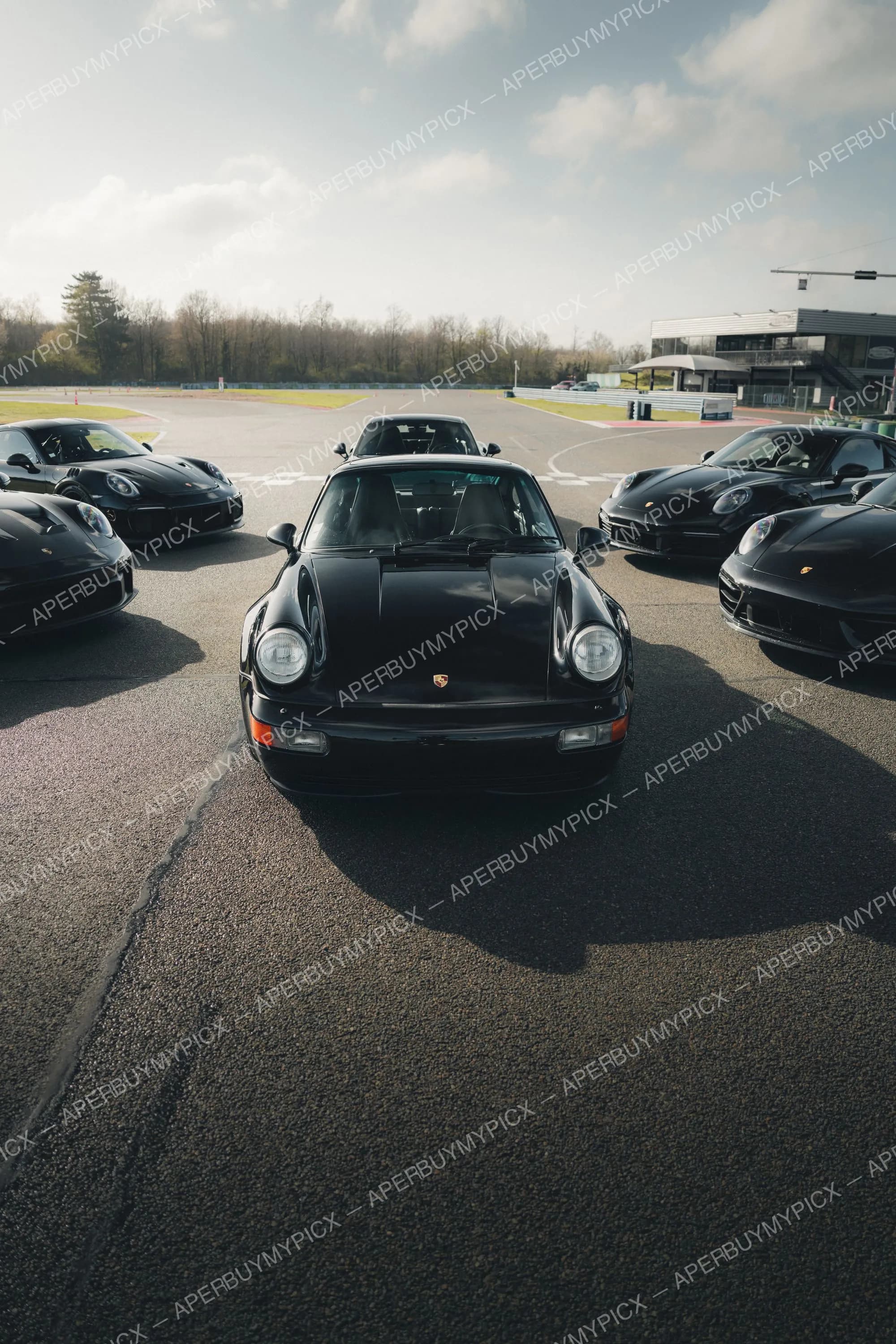 Porsche 911 993 noire vue de face au centre d'un circuit, entourée de plusieurs Porsche 911 noires, sous un ciel nuageux avec bâtiment et arbres en arrière-plan.
