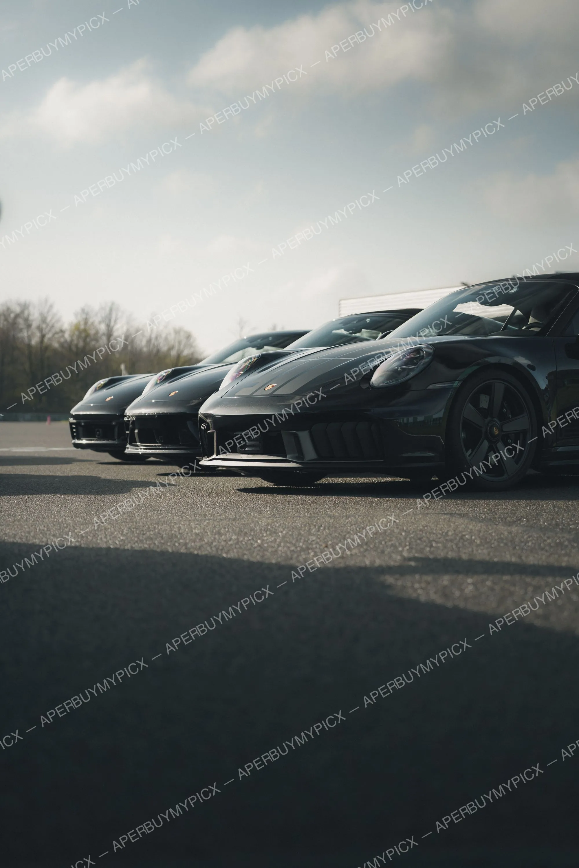 Trois Porsche 911 noires alignées sur un circuit, vue basse de face, sous un ciel nuageux avec arbres en arrière-plan