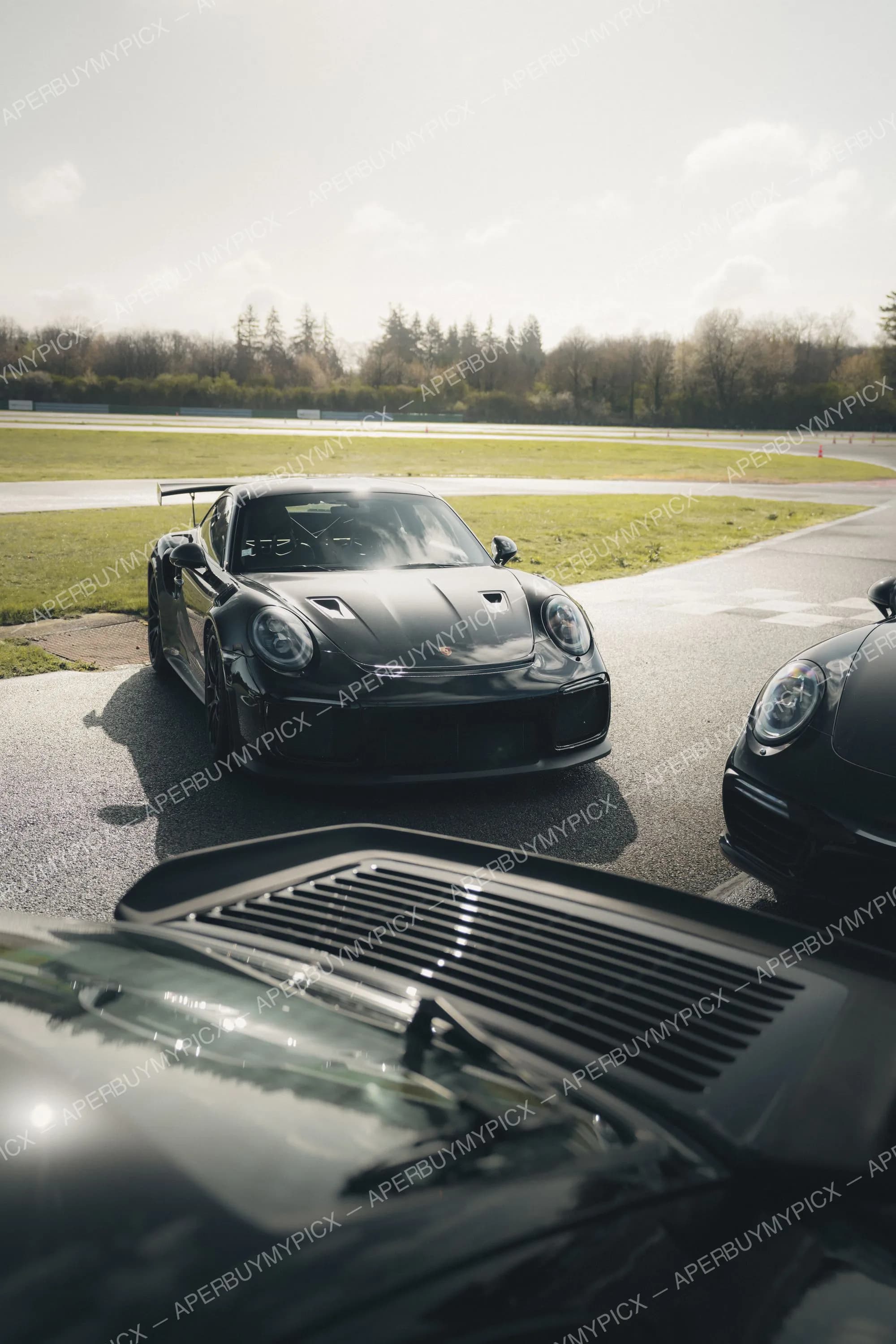 Porsche 911 GT3 RS noire garée sur un circuit ensoleillé, aileron arrière visible, à côté d'une autre Porsche 911 noire, avec piste, pelouse et arbres en arrière-plan.