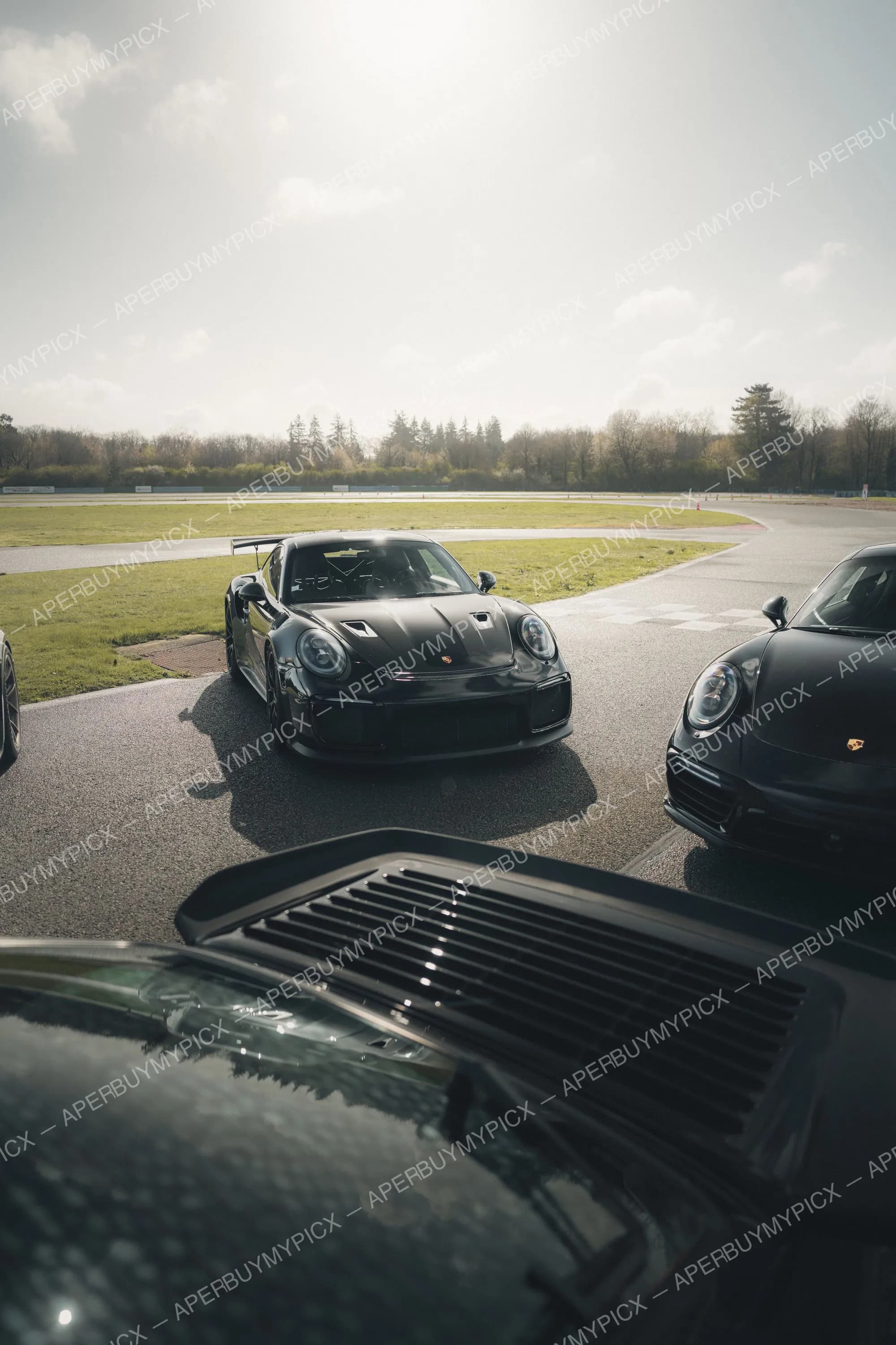 Porsche 911 GT2 RS noire garée en bord de piste par temps ensoleillé, avec une autre Porsche 911 noire à droite et un circuit bordé d'arbres en arrière-plan.