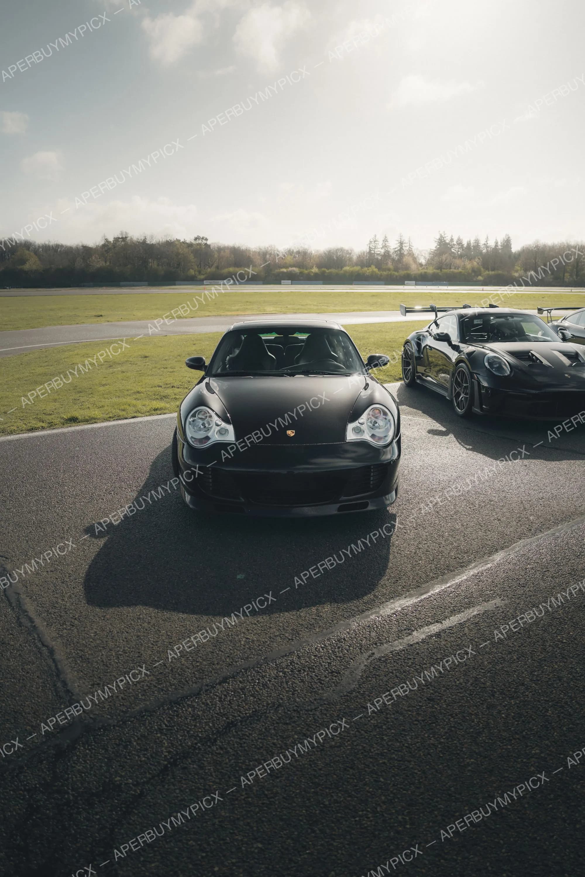 Deux Porsche 911 noires stationnées sur un circuit ensoleillé, une 996 Turbo au premier plan et une GT3 RS avec aileron à droite, herbe et arbres en arrière-plan.