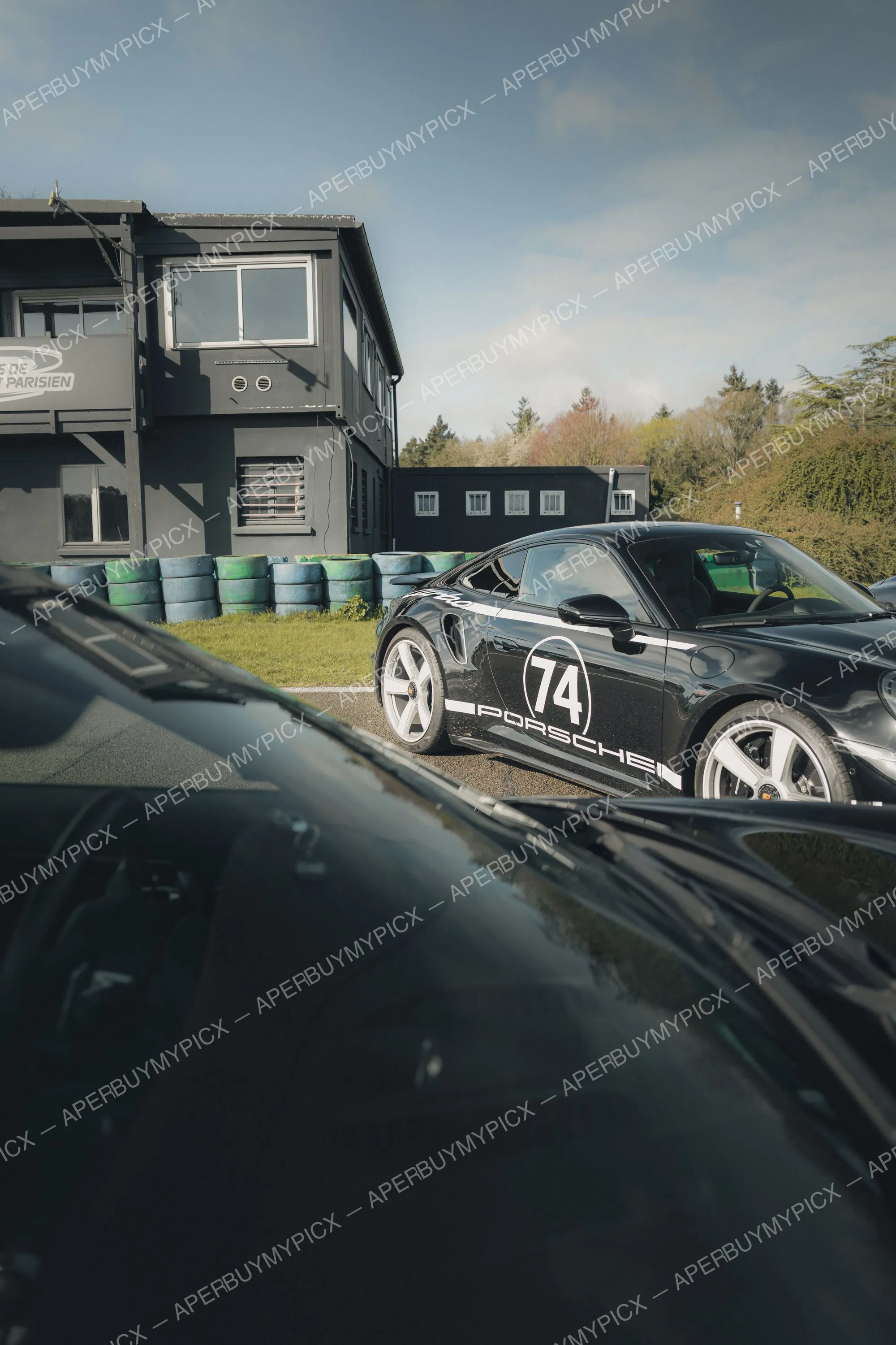 Porsche 911 Turbo noire portant le numéro 74 garée sur un circuit devant un bâtiment gris, avec des piles de pneus et une autre voiture noire au premier plan.