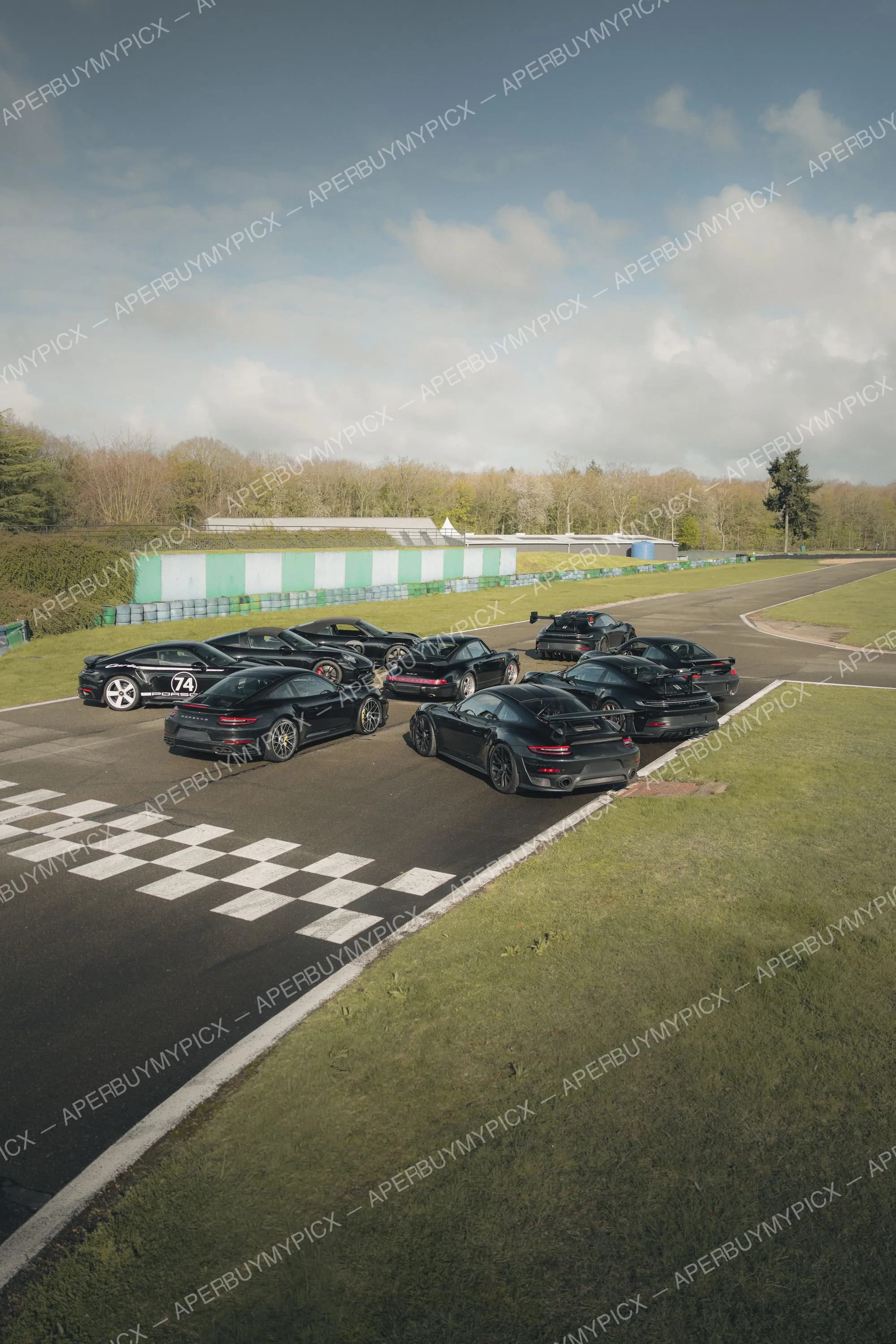 Groupe de Porsche 911 noires stationnées près de la ligne d'arrivée à damier d'un circuit, sous un ciel nuageux avec forêt en arrière-plan