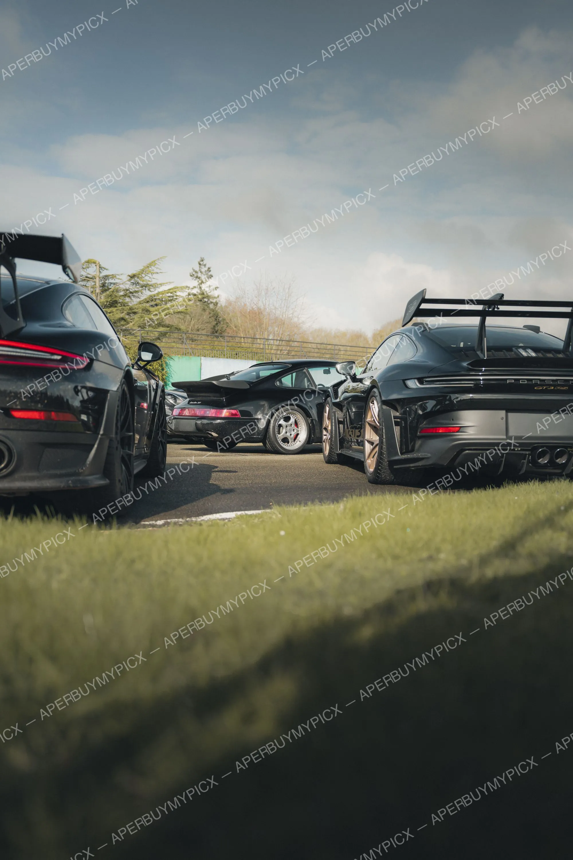 Trois Porsche 911 noires alignées en bord de piste, dont une GT3 RS avec aileron et jantes bronze, une 964 Turbo classique et une autre 911, sous un ciel nuageux avec pelouse au premier plan.