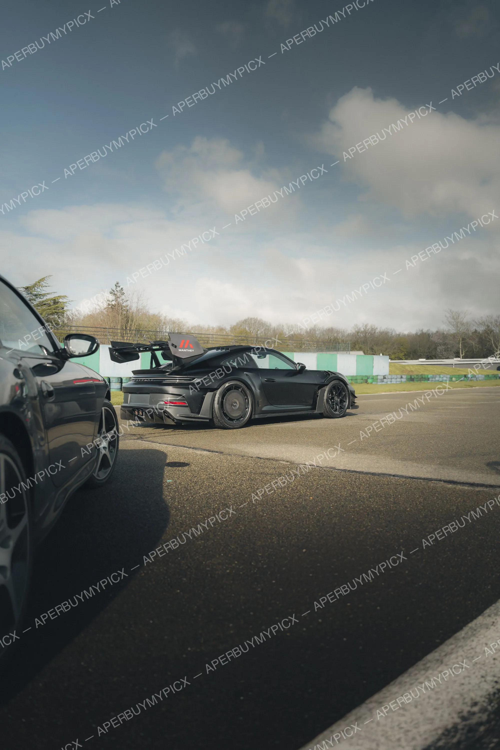 Porsche 911 GT3 RS noire avec kit Manthey et aileron arrière, garée sur un circuit sous un ciel nuageux, à côté d'une autre Porsche 911 sombre