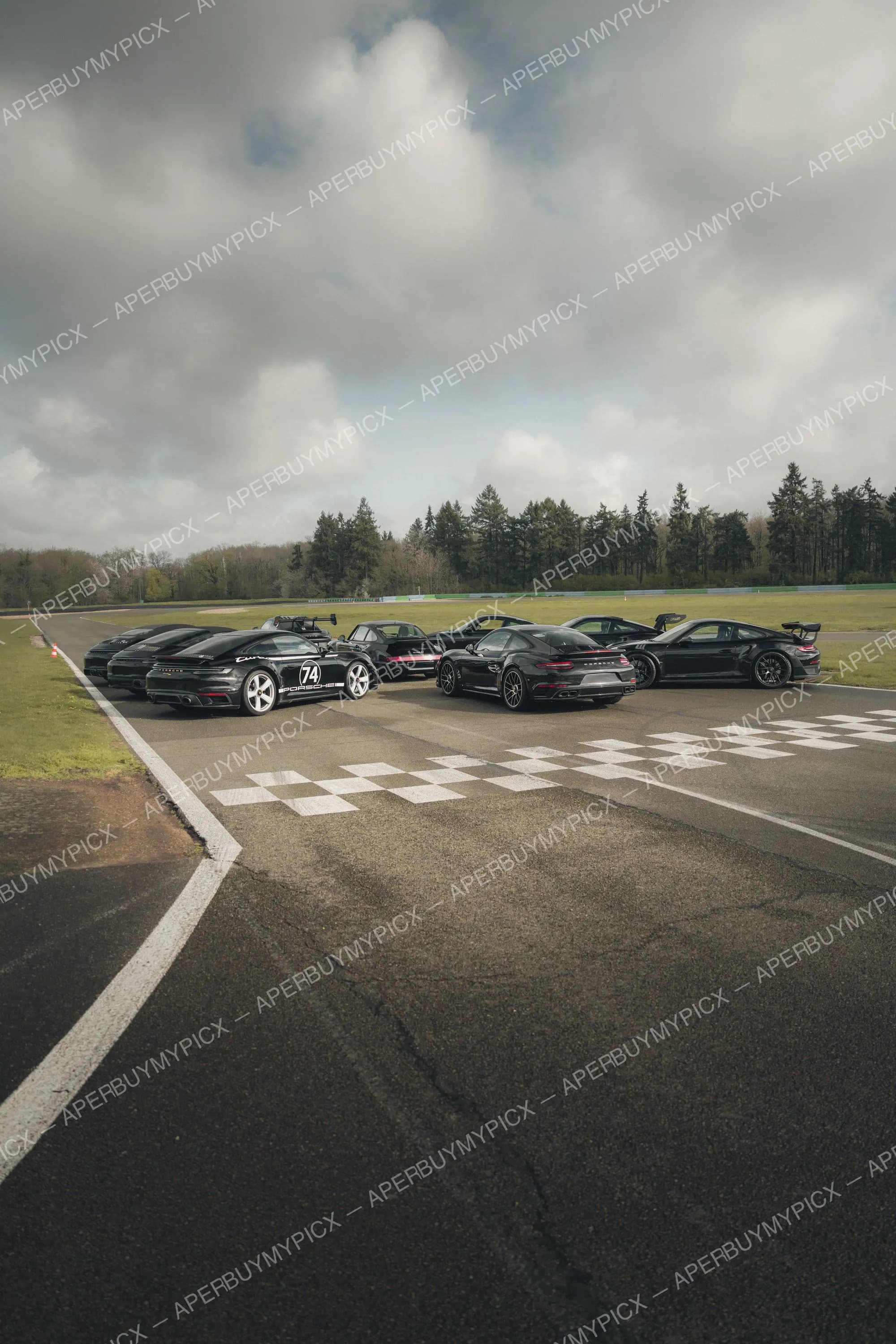 Groupe de Porsche 911 noires stationnées sur un circuit automobile près de la ligne de départ à damier, sous un ciel nuageux avec des arbres en arrière-plan